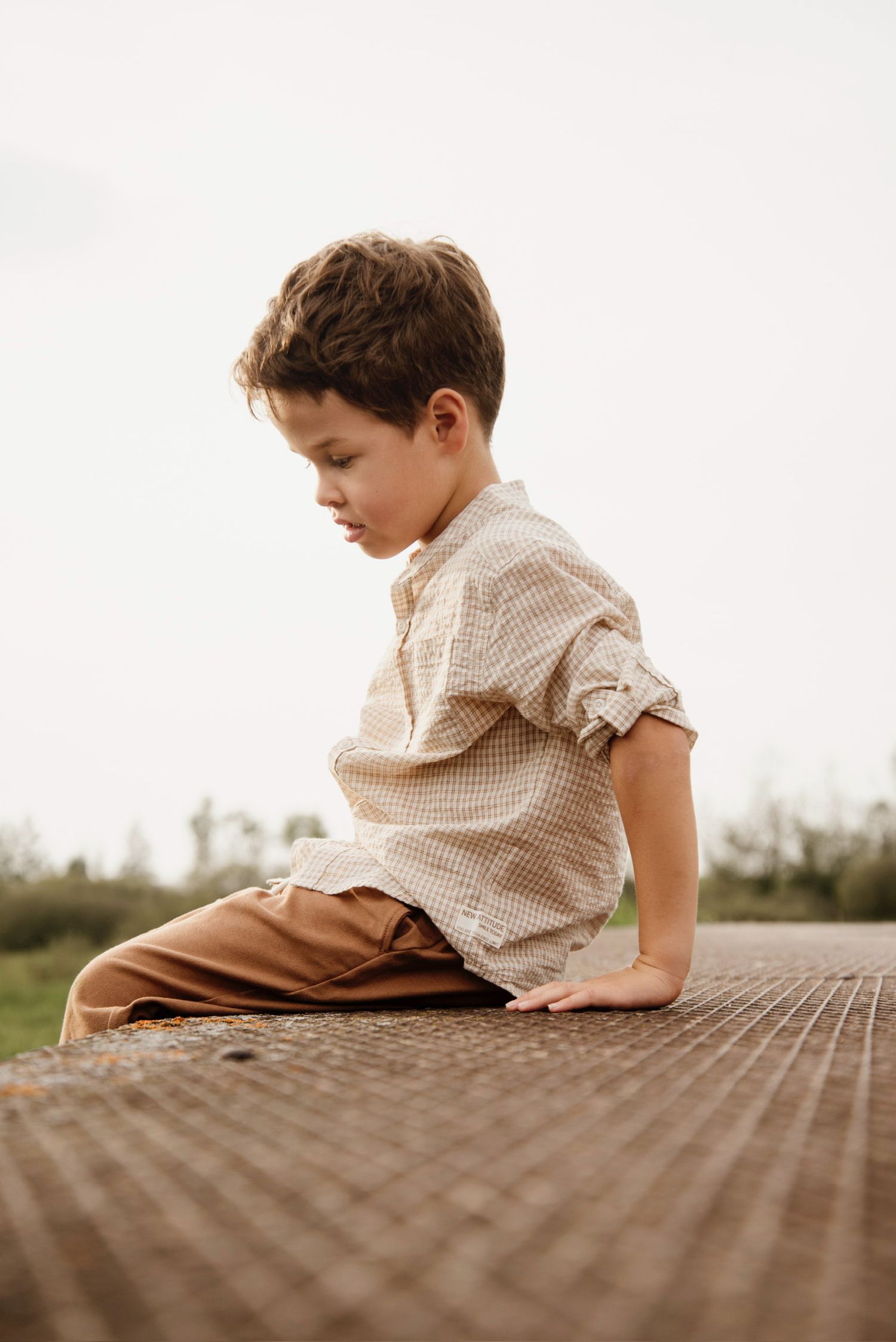 Young boy in beige shirt and tan pants sitting on wooden deck, looking down thoughtfully outdoors.