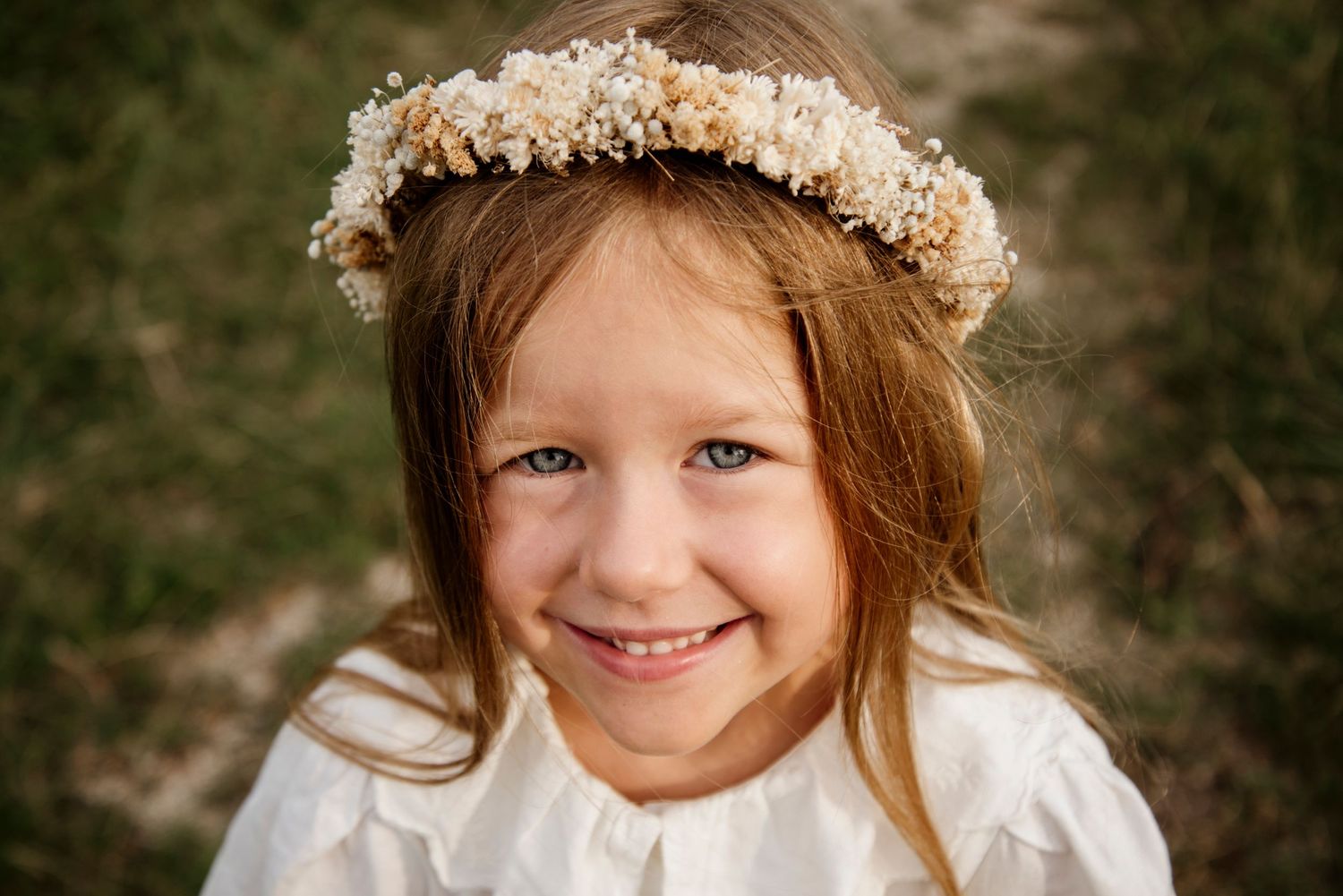Smiling young girl wearing a floral crown and white dress, photographed outdoors in a charming portrait.