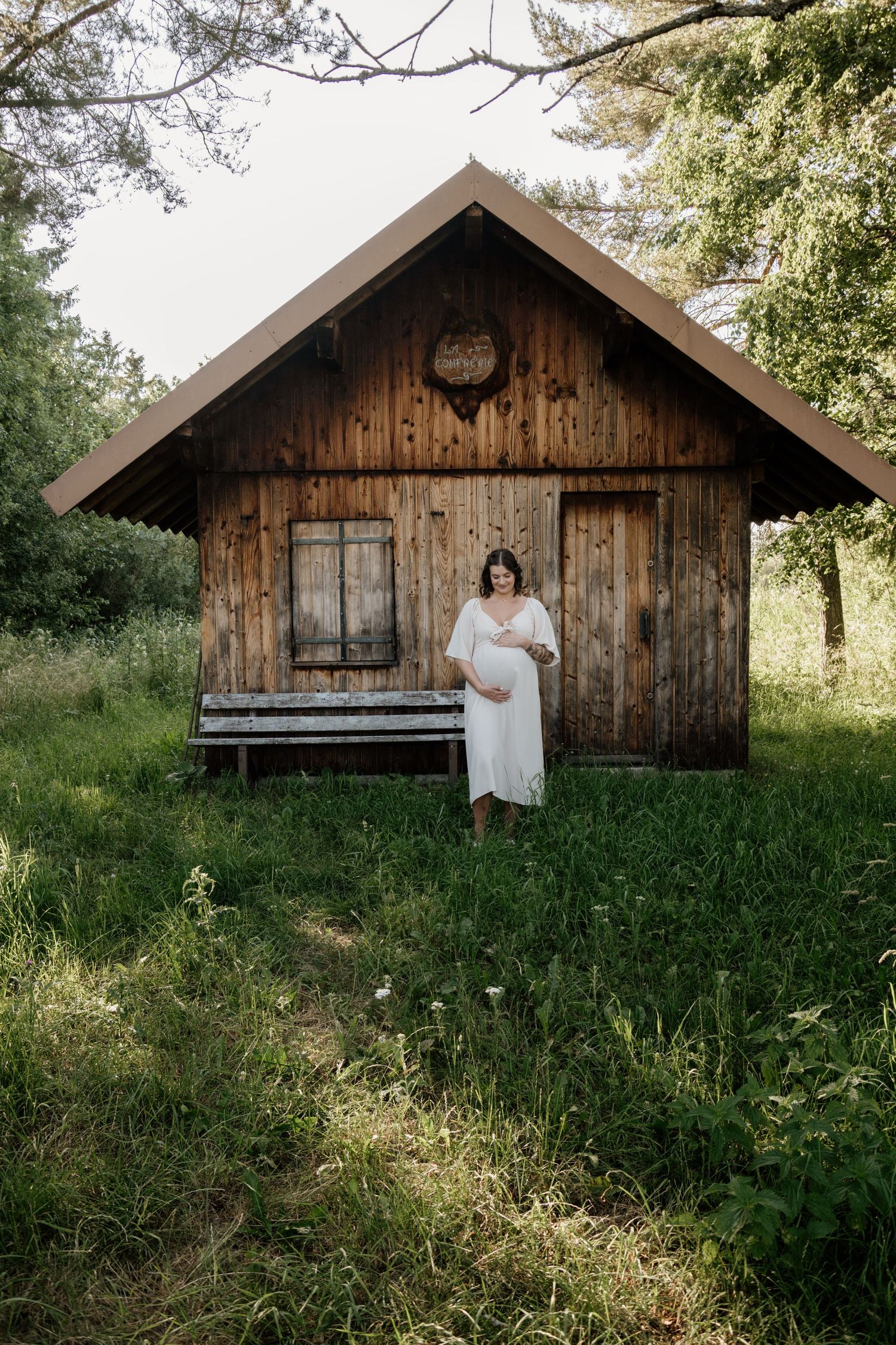 Pregnant woman in white dress stands before rustic wooden cabin surrounded by greenery.