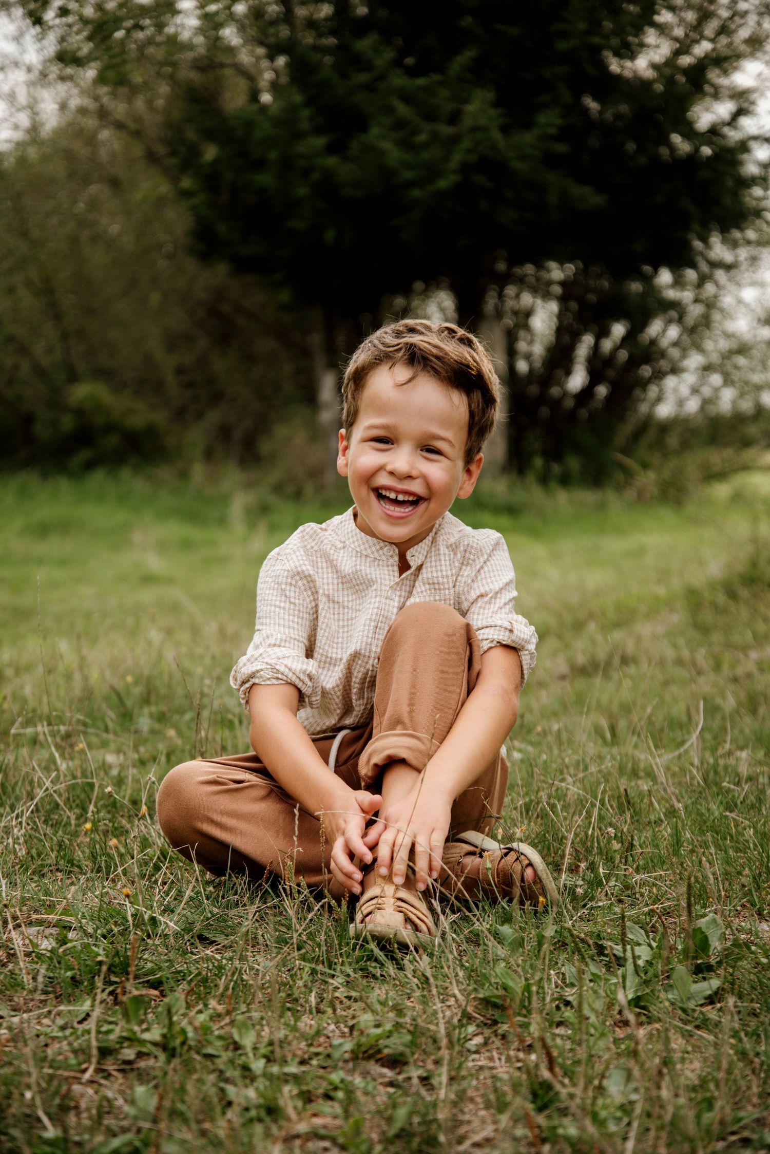 A joyful young boy sits cross-legged on green grass, laughing brightly, wearing a beige shirt and tan pants outdoors.