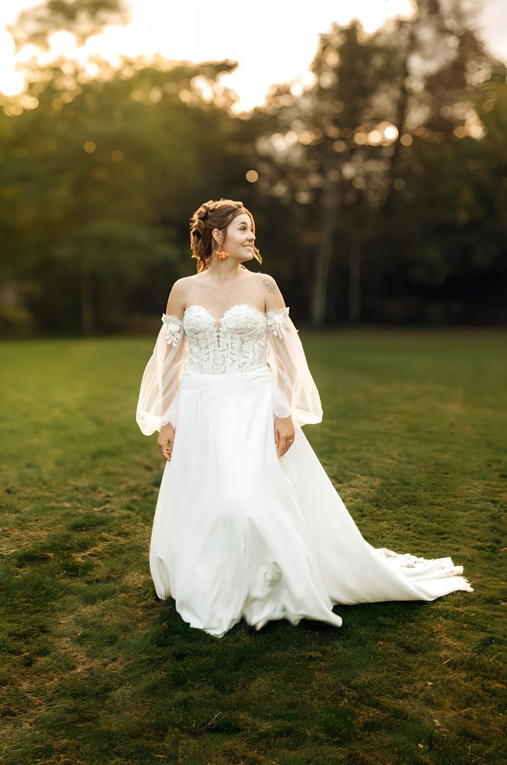 Elegant bride wearing off-shoulder white wedding dress with bishop sleeves stands on grass during sunset.