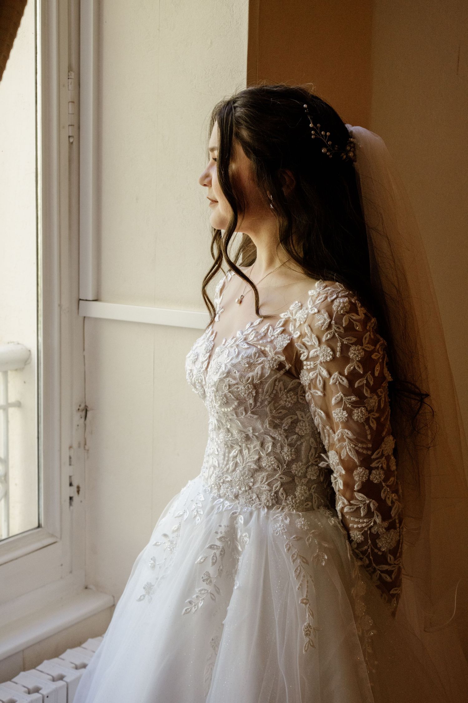 Bride in a long-sleeve lace wedding gown gazes out a sunlit window on her wedding day.