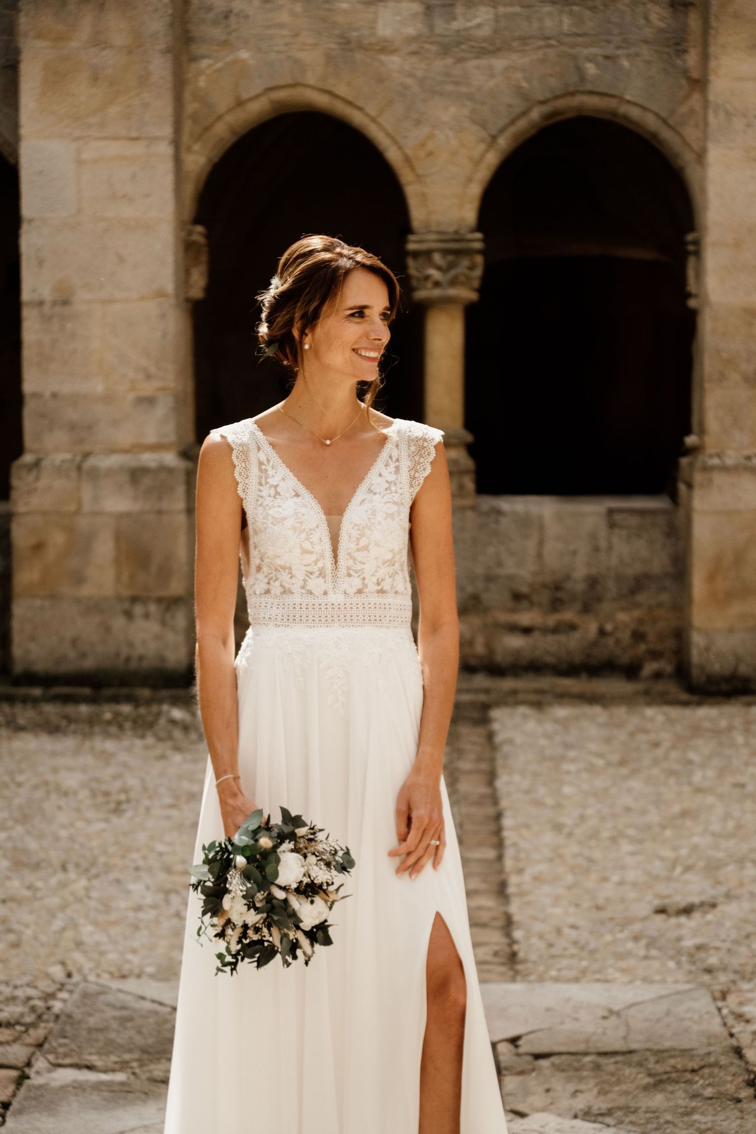 Smiling bride in V-neck lace wedding dress holds white bouquet before stone arched building in sunlight.