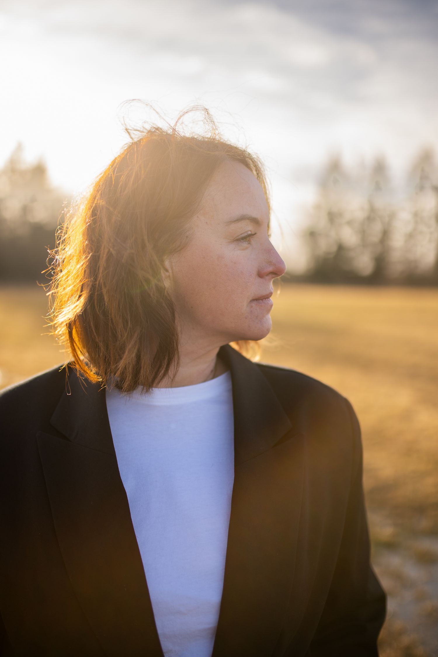 Woman in black blazer and white shirt stands in golden sunlight outdoors, with trees and open field in background.