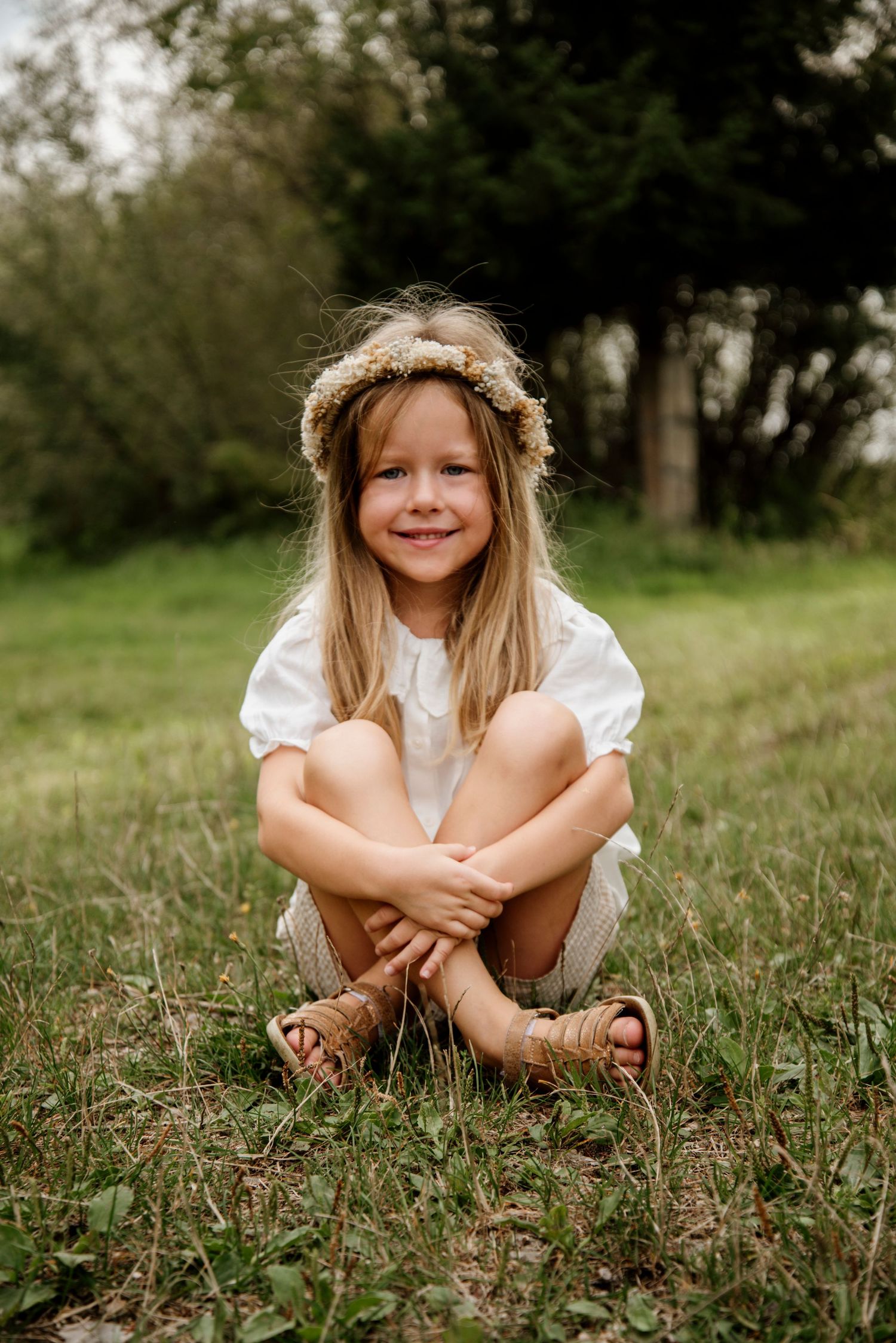 A smiling young girl with a floral crown sits cross-legged on grass in a white dress and sandals, surrounded by trees.