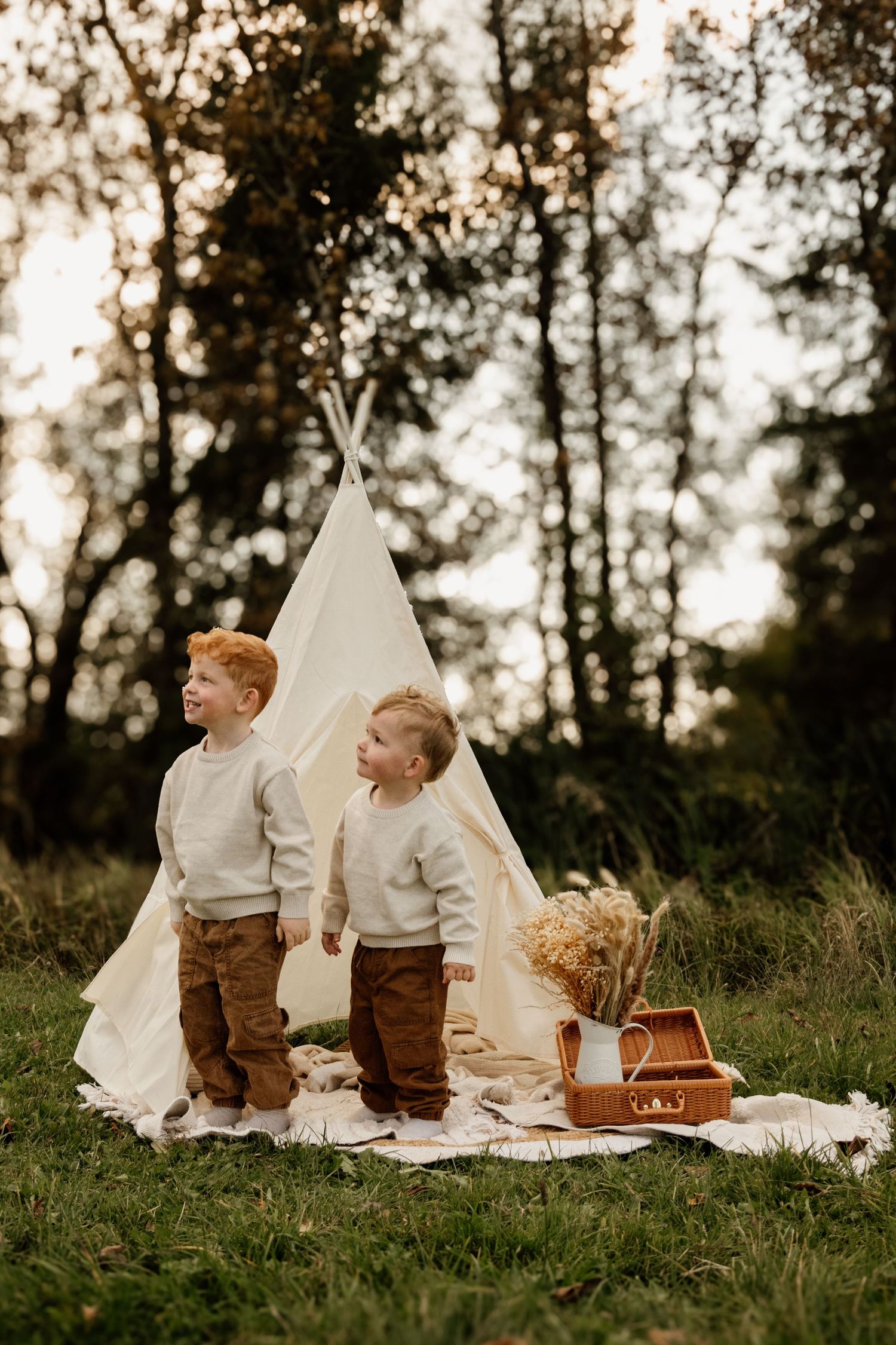 Two young redhead brothers in cream sweaters standing before a teepee tent in an autumn outdoor setting.
