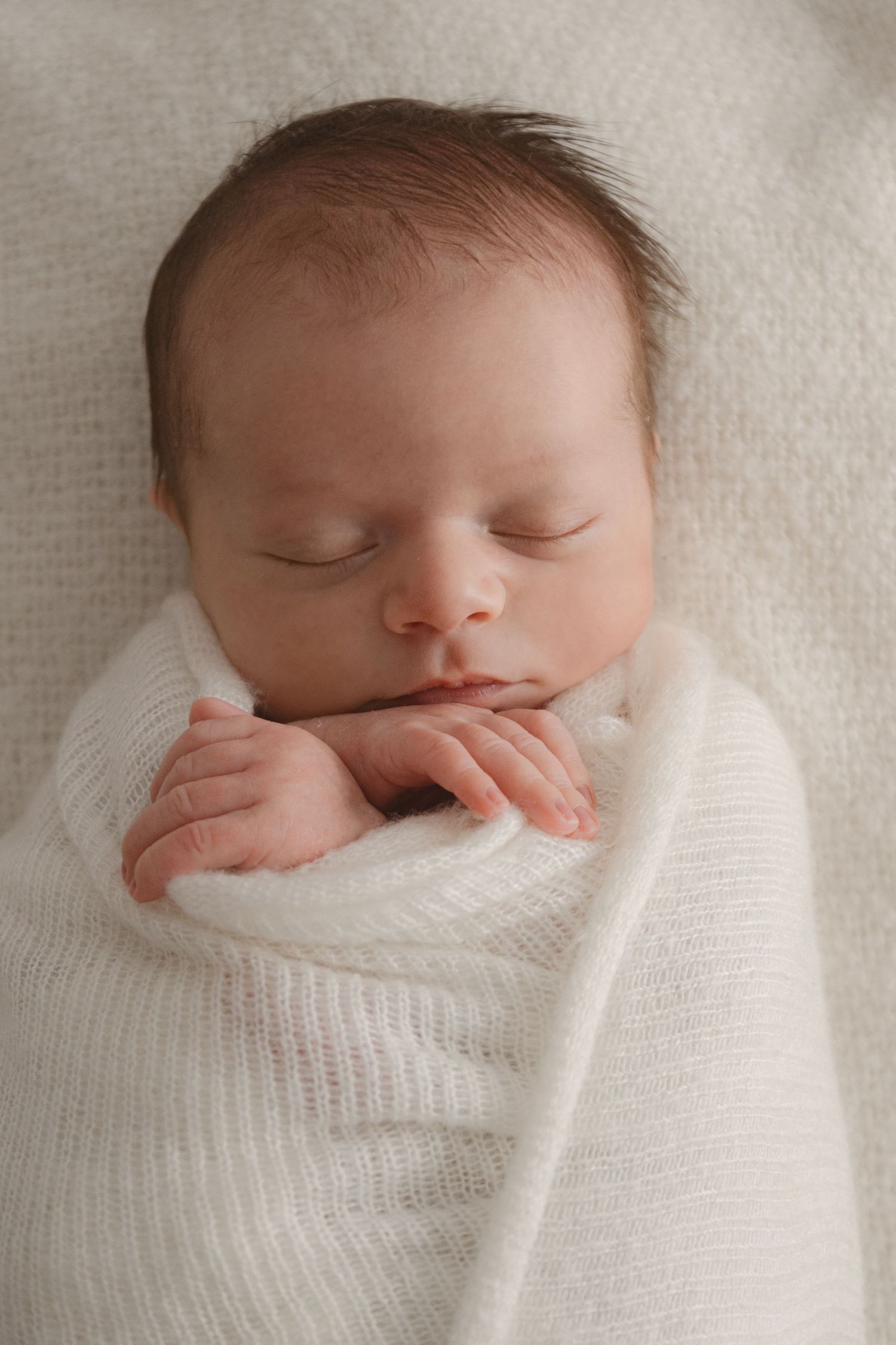 Peaceful newborn baby sleeping swaddled in a soft white wrap with tiny hands visible under chin.