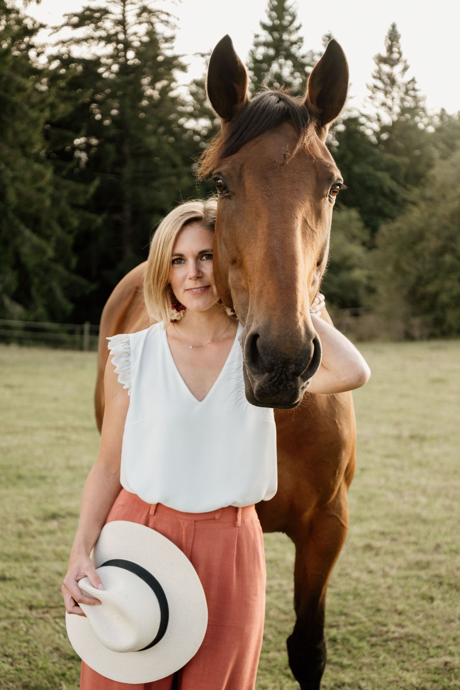 A blonde woman in a white top and pink skirt stands beside a brown horse in a green field.