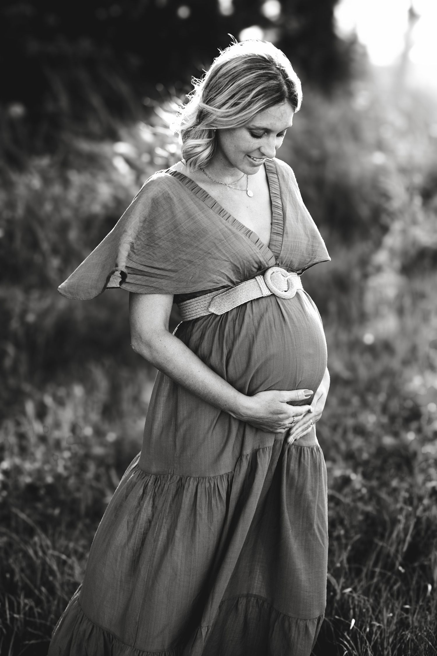 Black and white photo of pregnant woman in belted dress cradling baby bump in field.