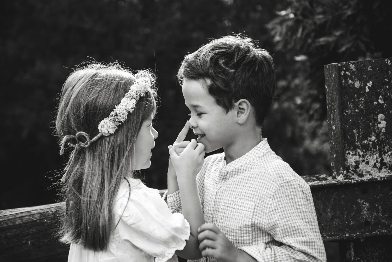 Black and white photo of two young children sharing a tender moment outdoors, girl wearing floral crown.