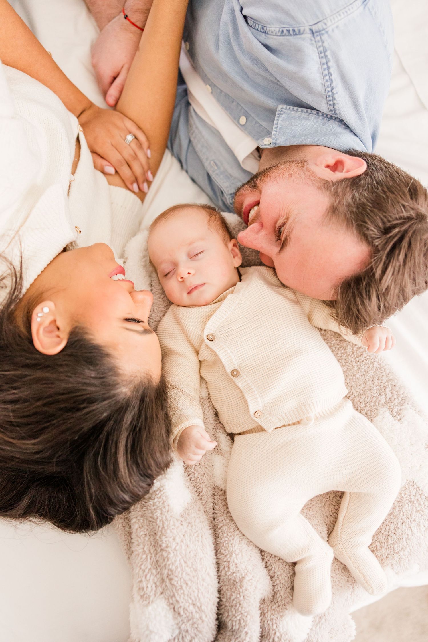 A baby in white sleepwear sleeps peacefully on a soft blanket while parents snuggle close on either side.