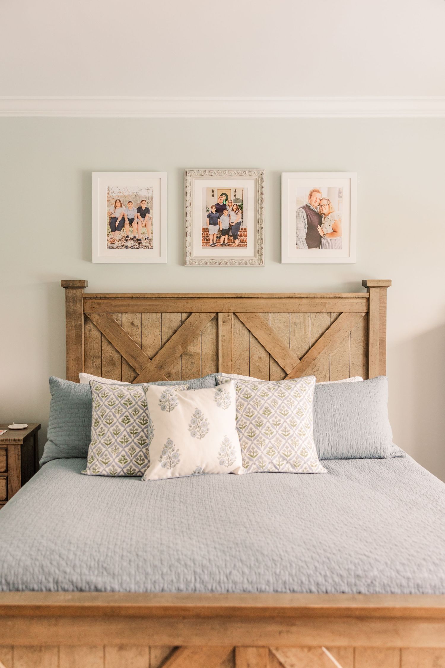 A rustic farmhouse bedroom with barn door style wooden headboard, gray bedding, decorative pillows and framed photos above.