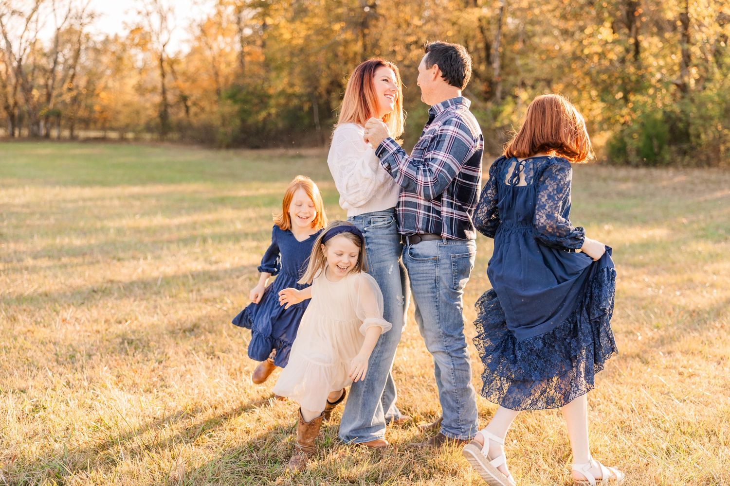 A family in coordinating navy and white outfits stands together in a golden autumn field at sunset.