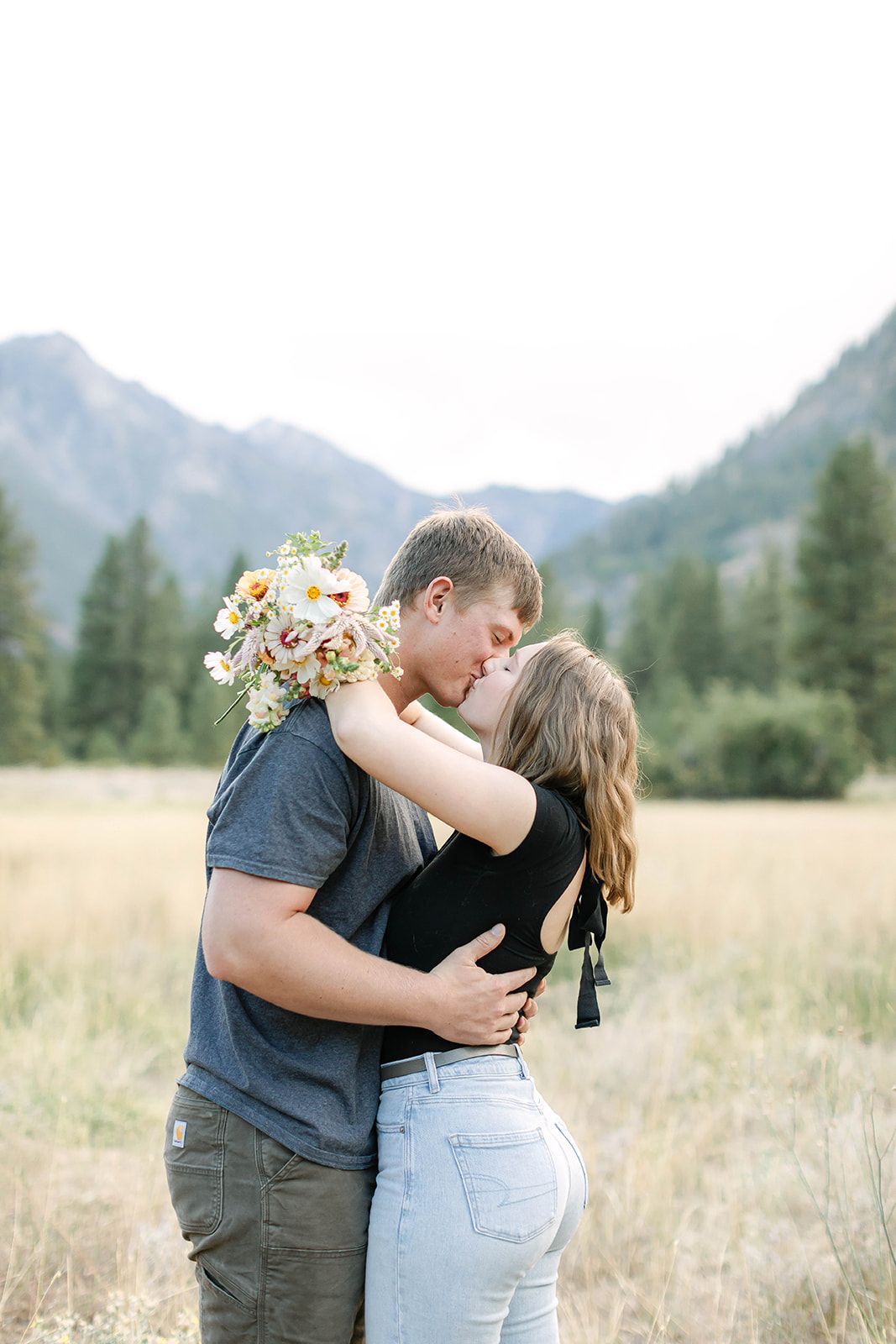 Romantic couple embracing in a sunny meadow with mountains and evergreen trees in the distance.