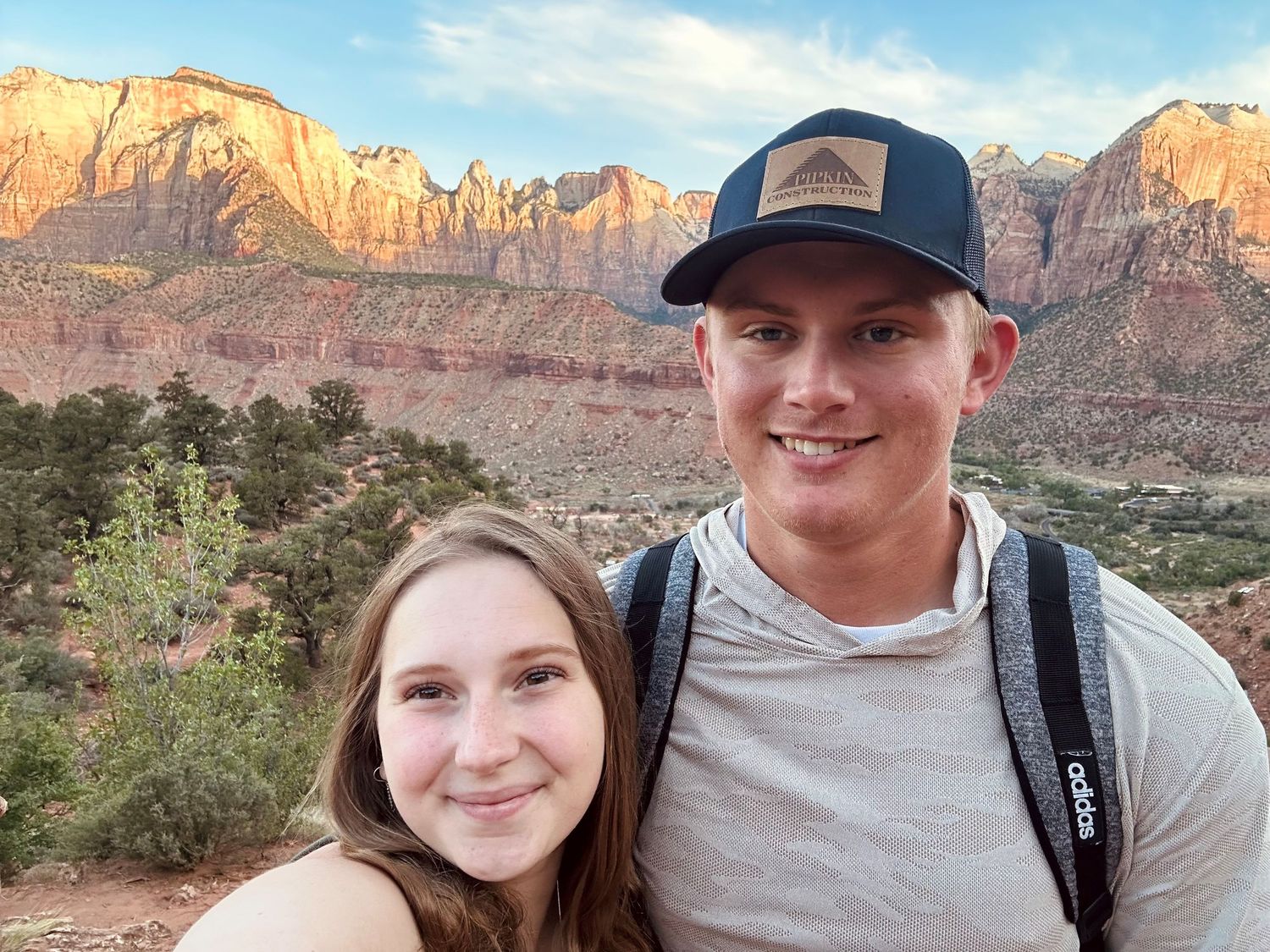Two people take a selfie with red rock cliffs of Zion National Park in the background.