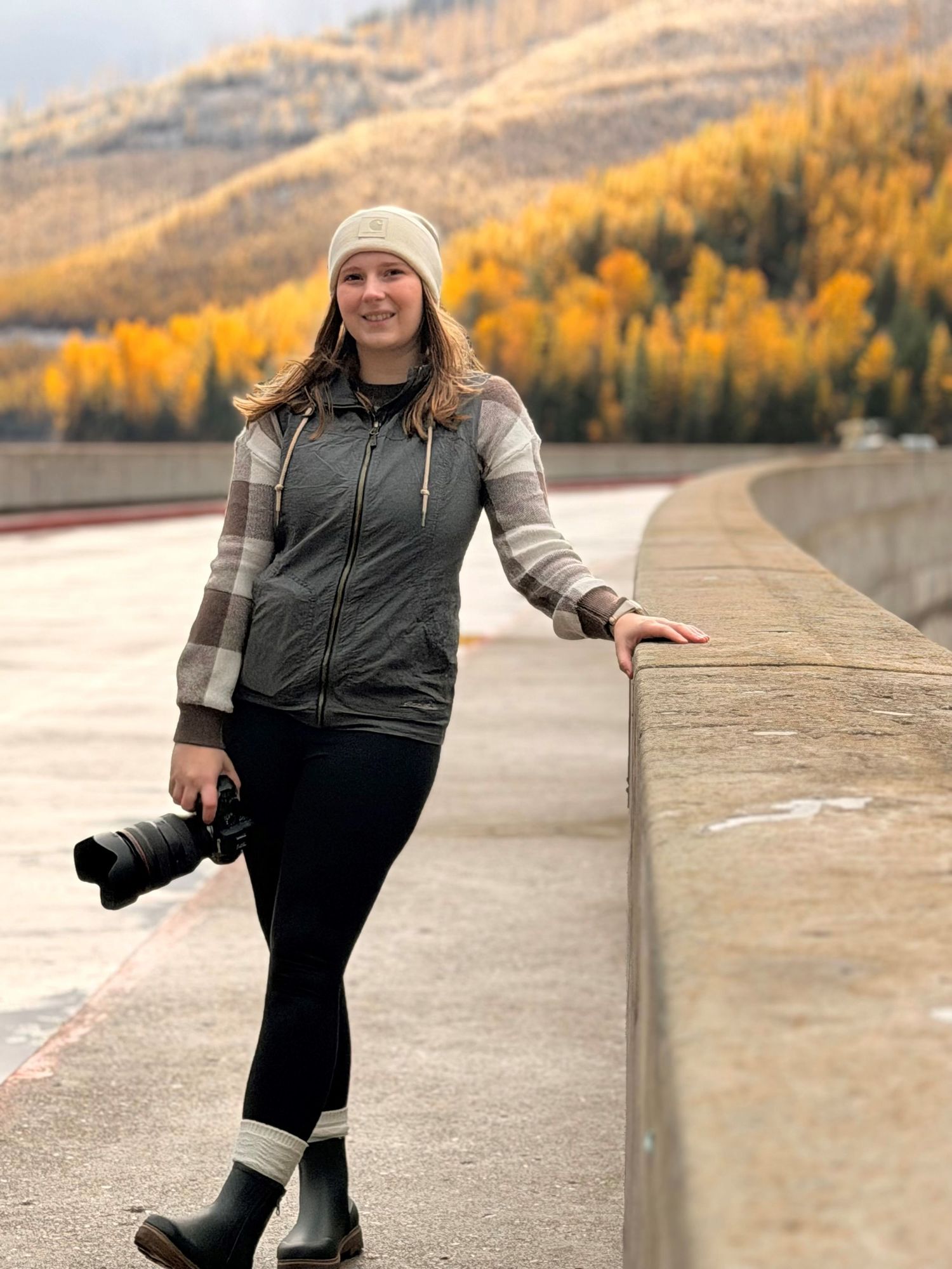 A person in winter attire poses along a concrete wall with autumn foliage and mountains in the background.