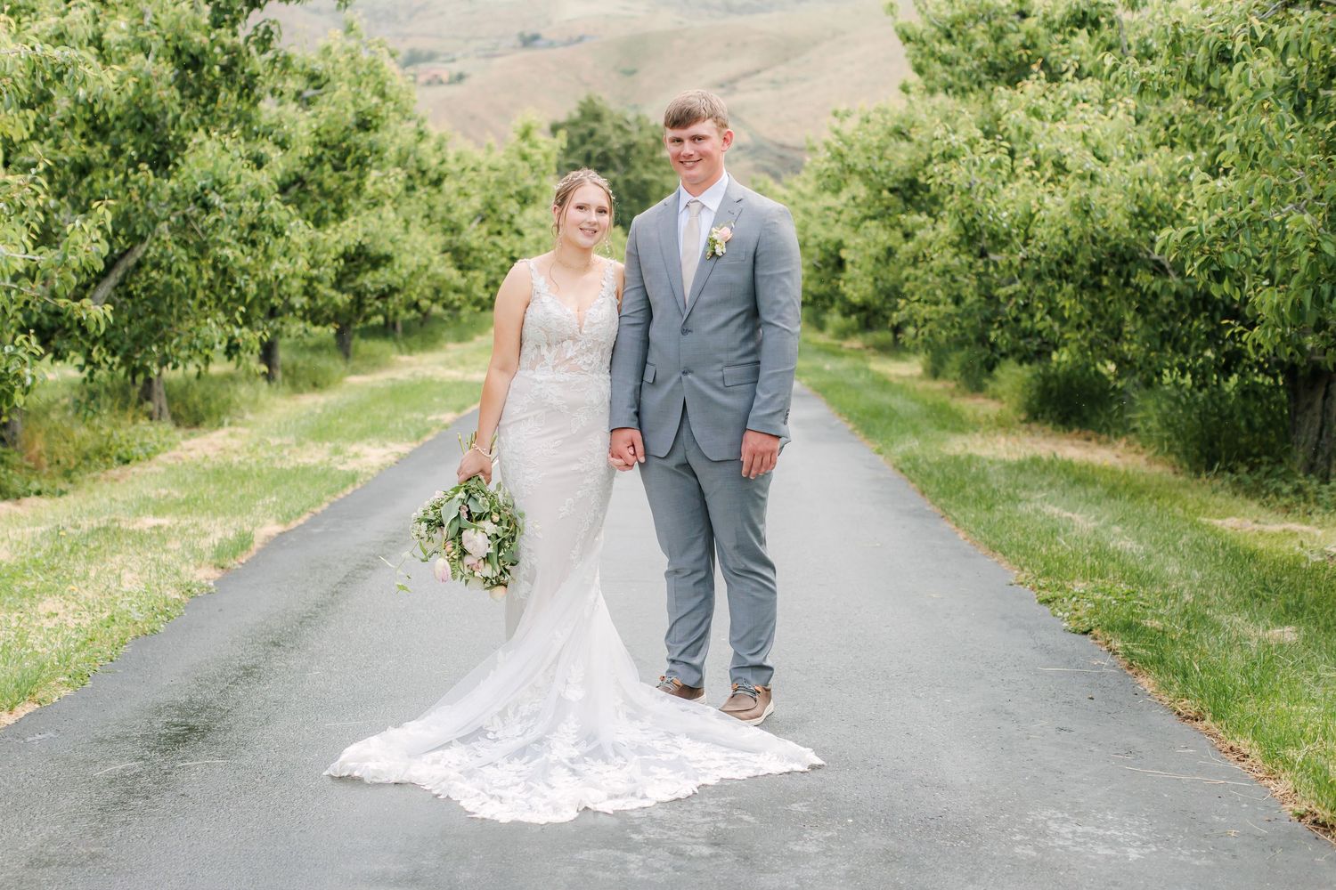 Couple walking down a tree-lined path in formal wedding attire with mountains in the background.