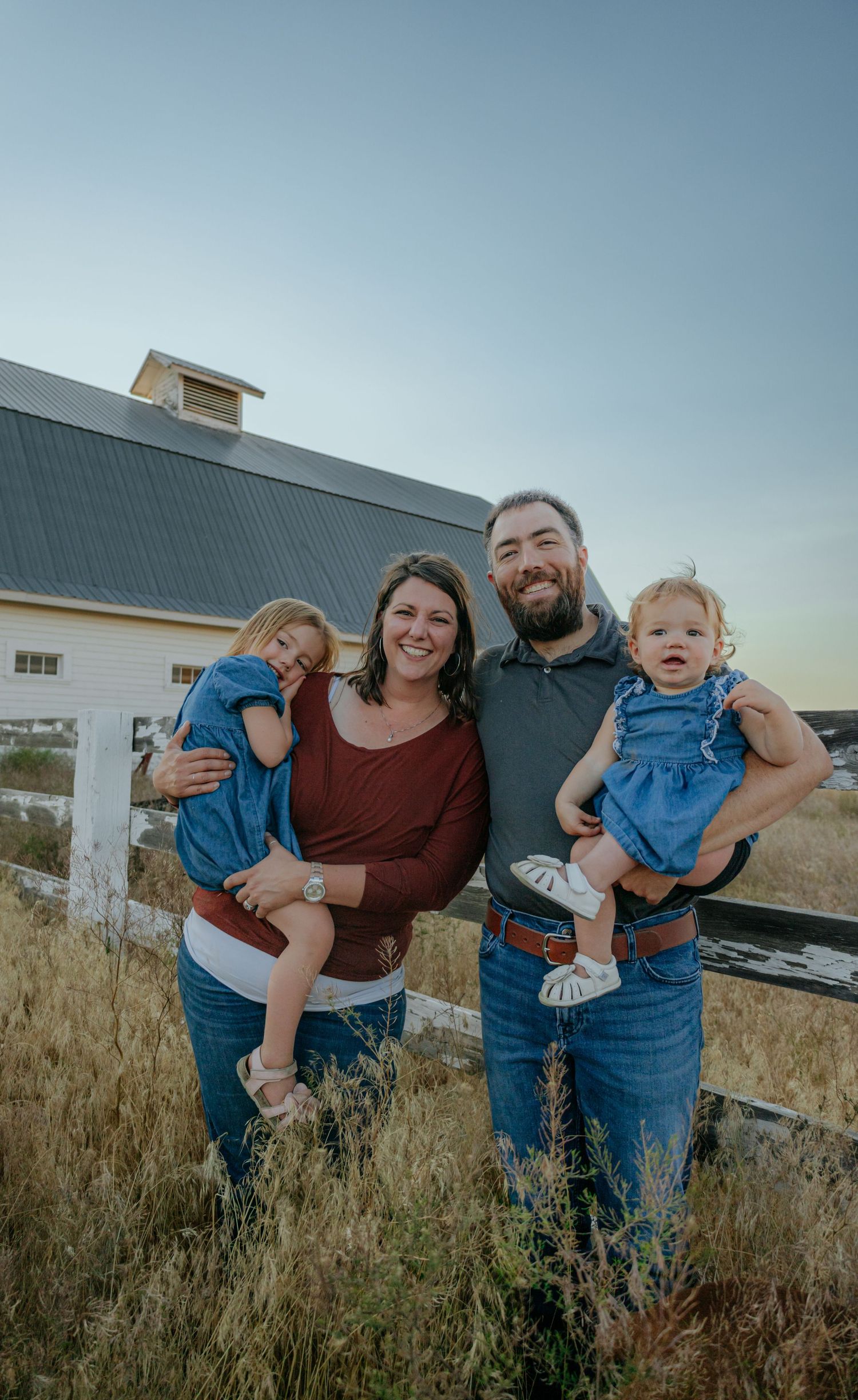Casual family portrait with white barn and countryside backdrop at sunset.