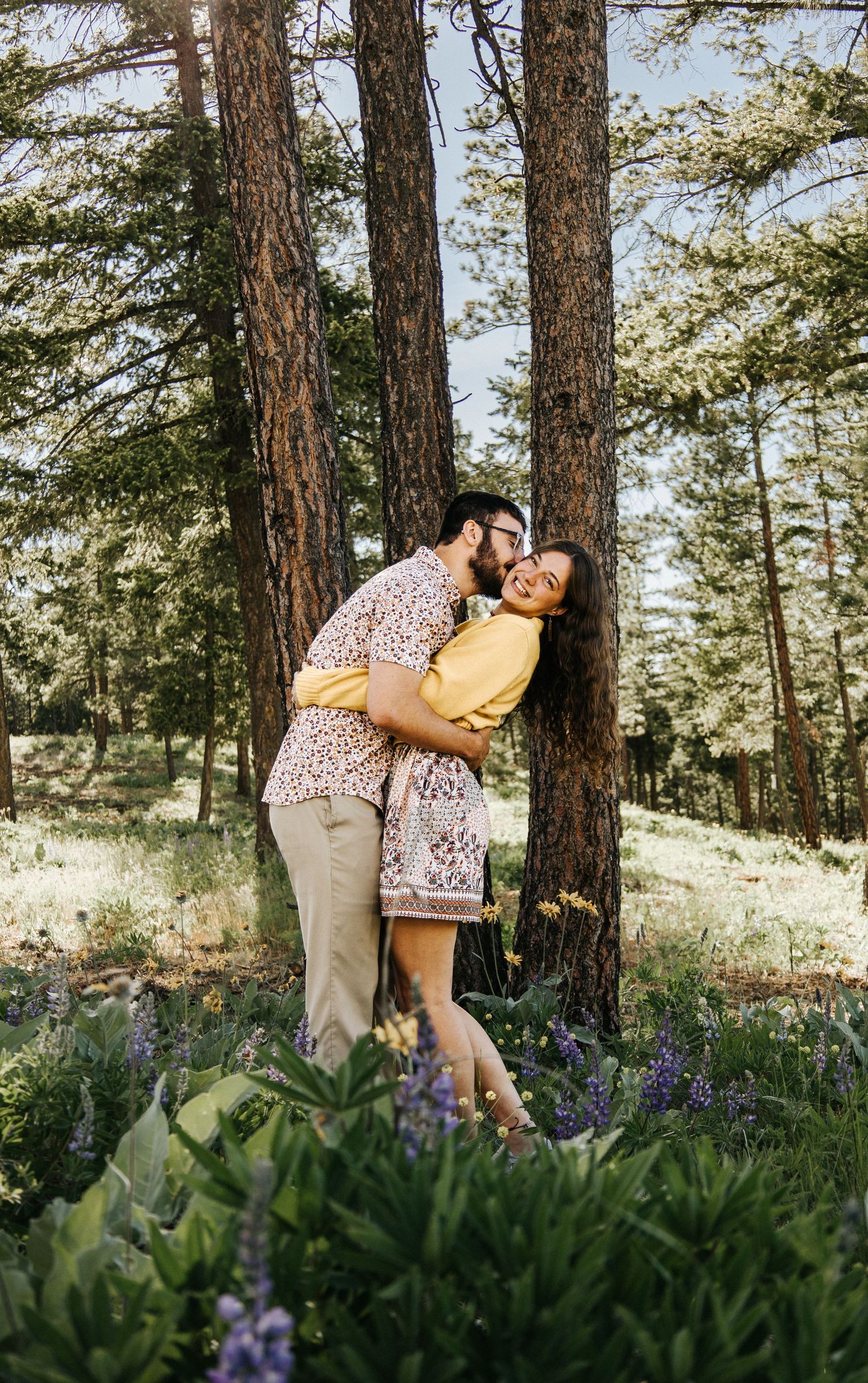Romantic couple poses in a meadow of purple lupines with tall pine trees in the background.