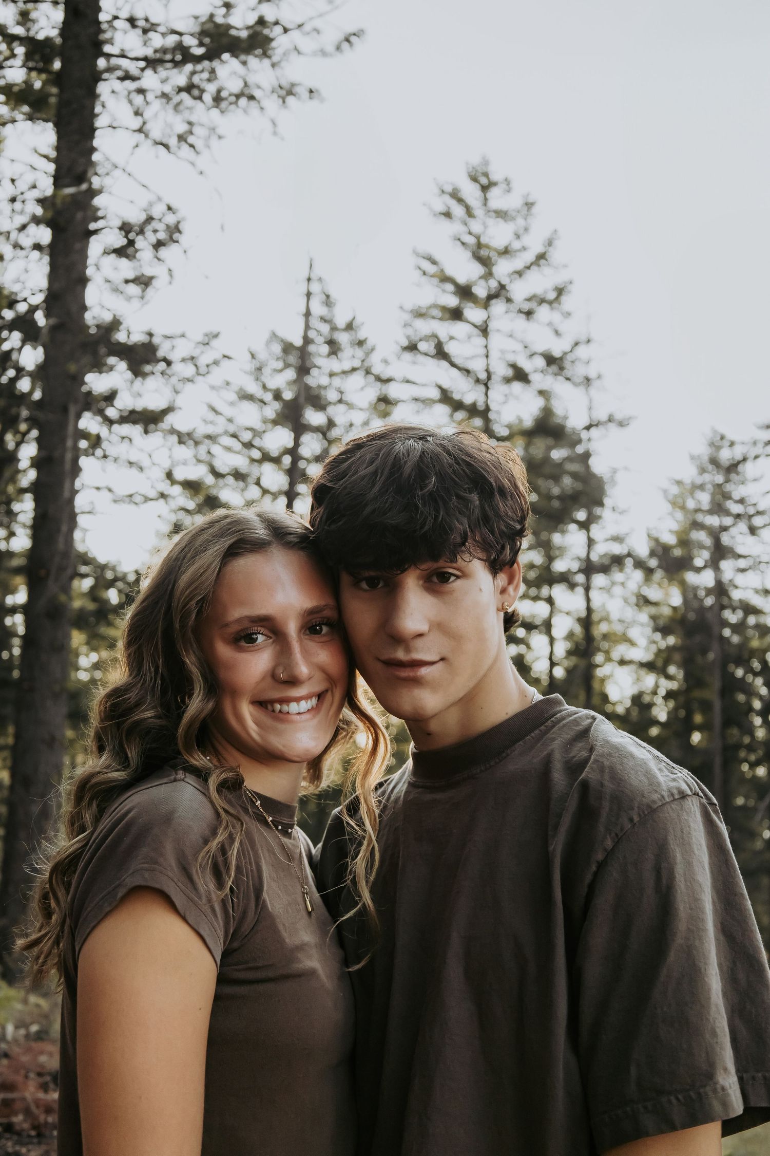 Two people pose together in a forest setting with evergreen trees in the background.