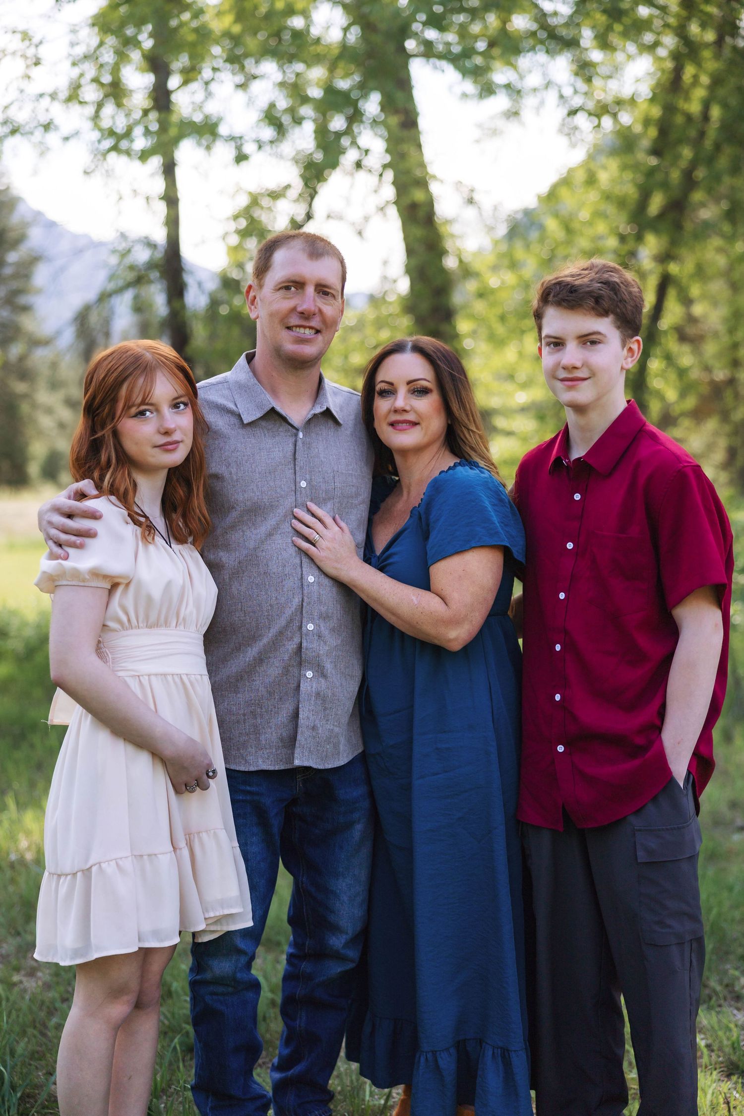 Family group stands together in coordinated outfits against a natural outdoor backdrop.