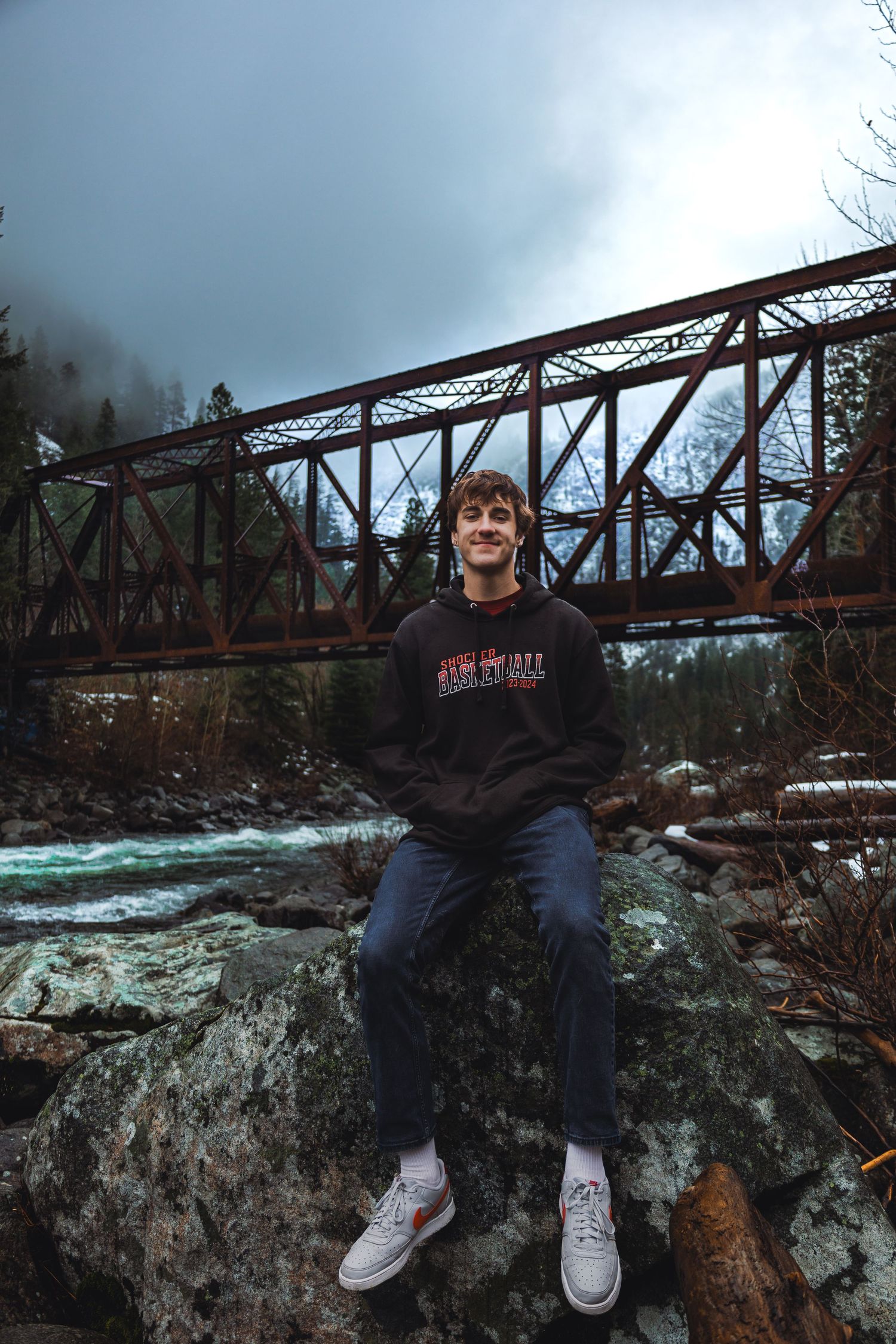 Person sits on rocky outcrop near rushing river with rustic bridge in background.
