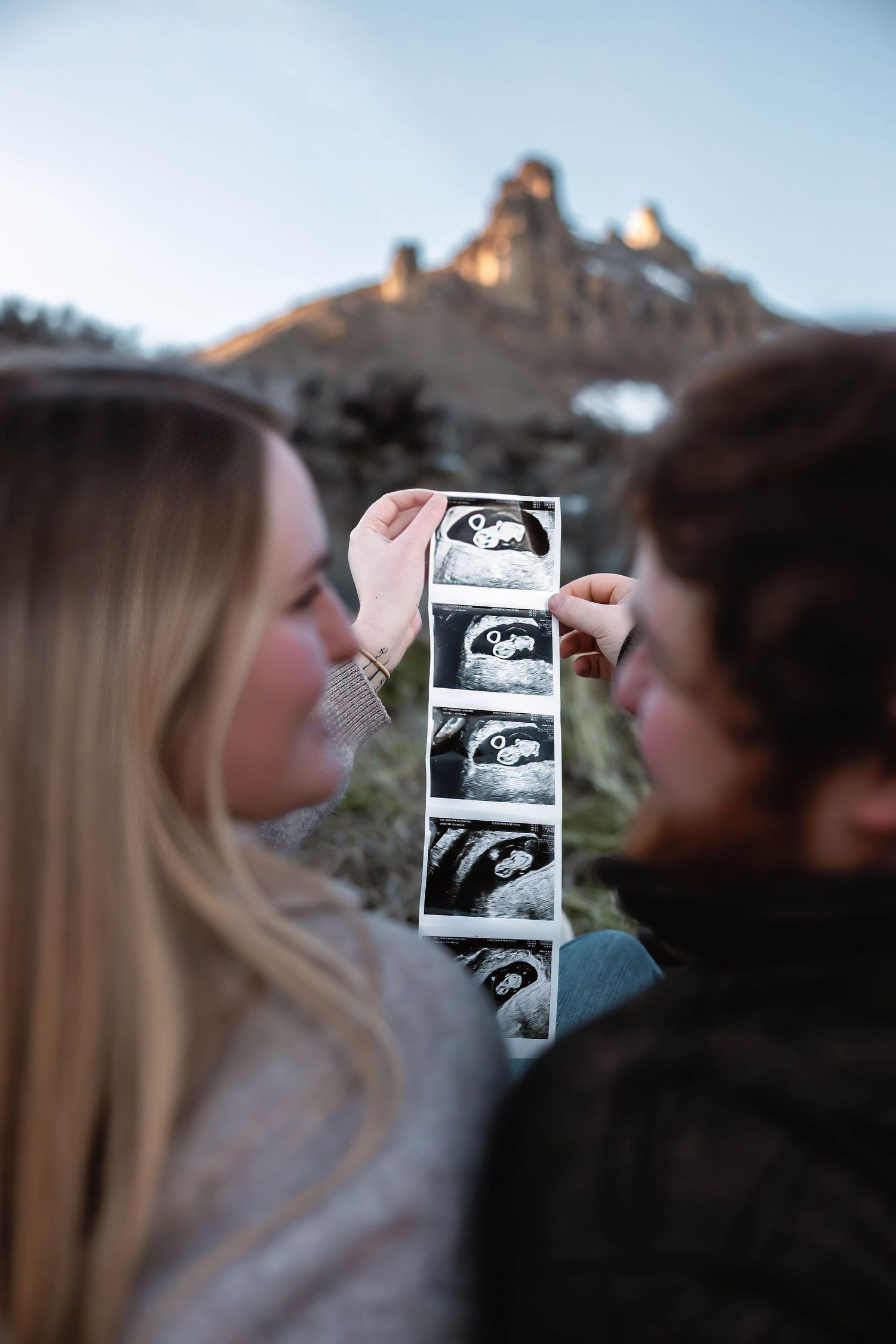 Couple holds up ultrasound photos outdoors during sunset with mountain backdrop.