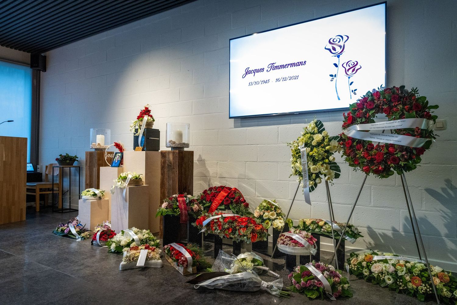 A funeral memorial display with floral wreaths, bouquets, and a screen showing a tribute name and dates in a modern hall.