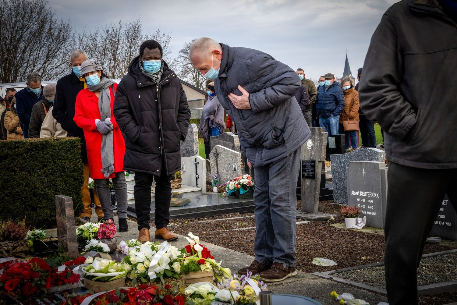 People paying respects at a graveside funeral ceremony with floral tributes in a cemetery.
