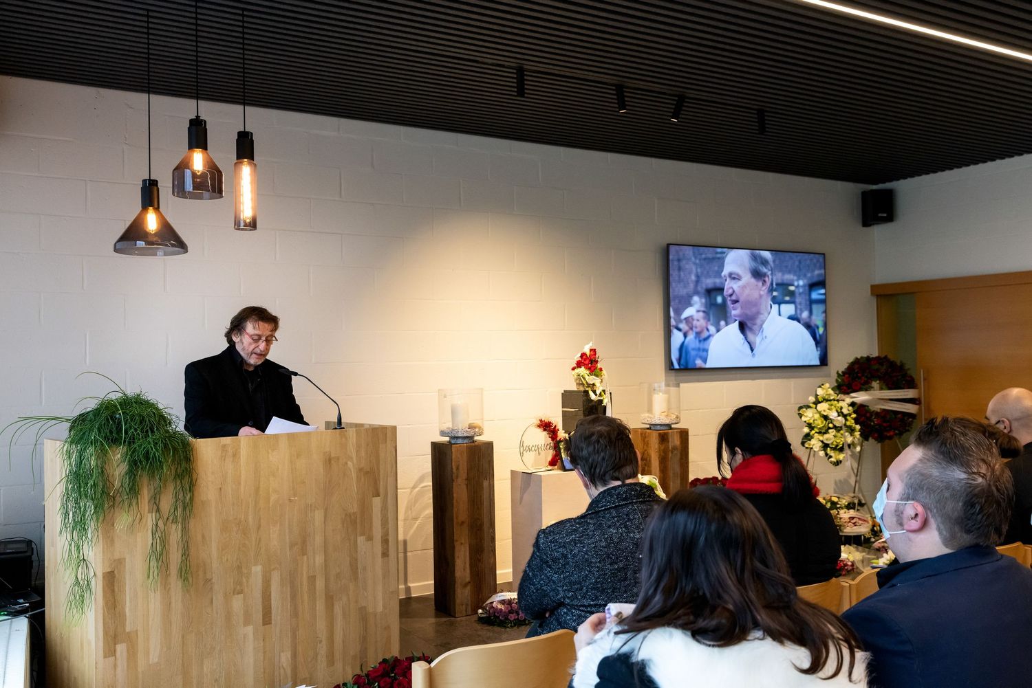 Speaker at podium addresses seated audience in modern venue with TV screen displaying presentation in background.