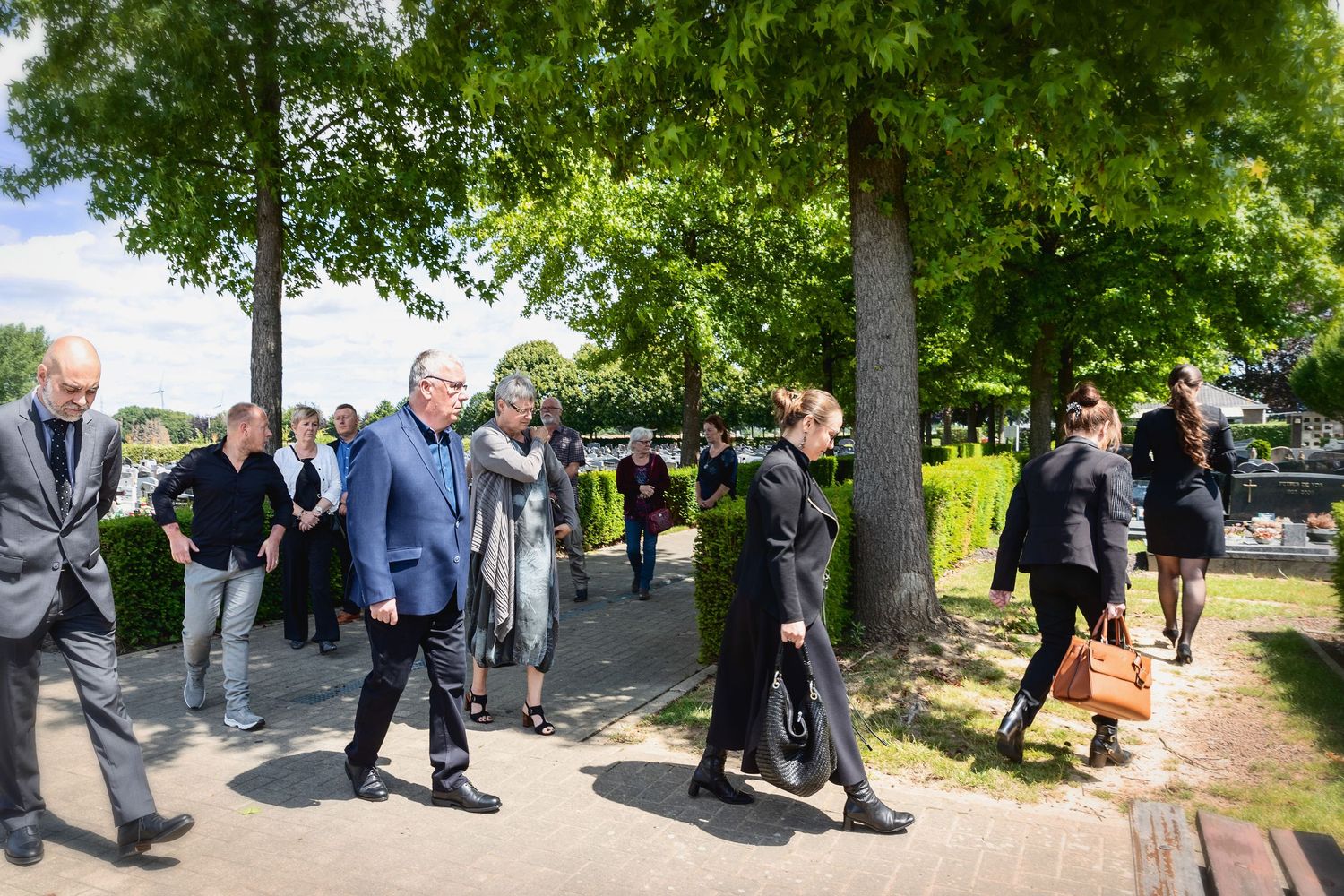 Group of people dressed in black walking along a tree-lined path, likely attending a formal outdoor gathering or funeral.