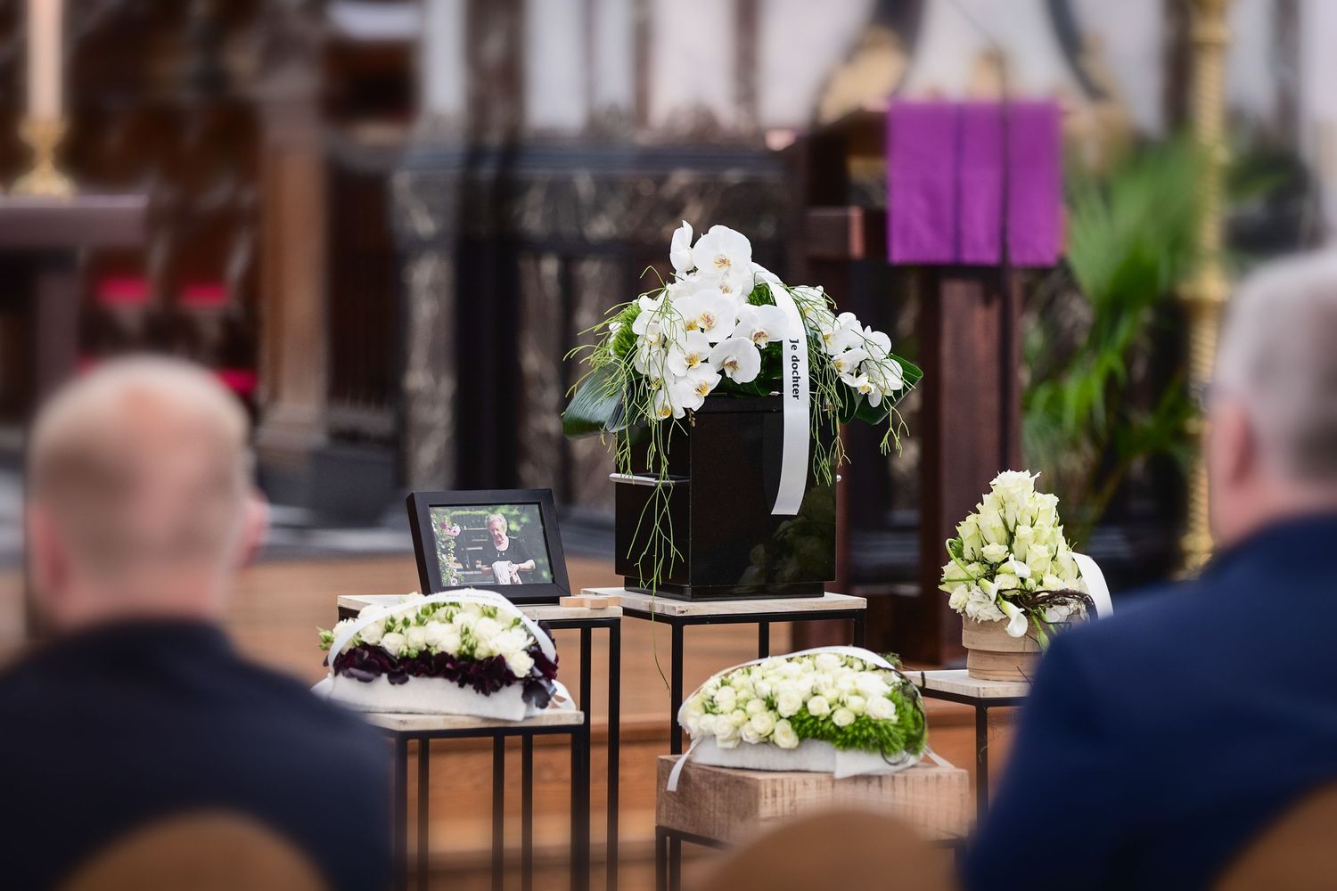 Funeral service in a church with white floral arrangements, an urn, and a framed photo on display tables.