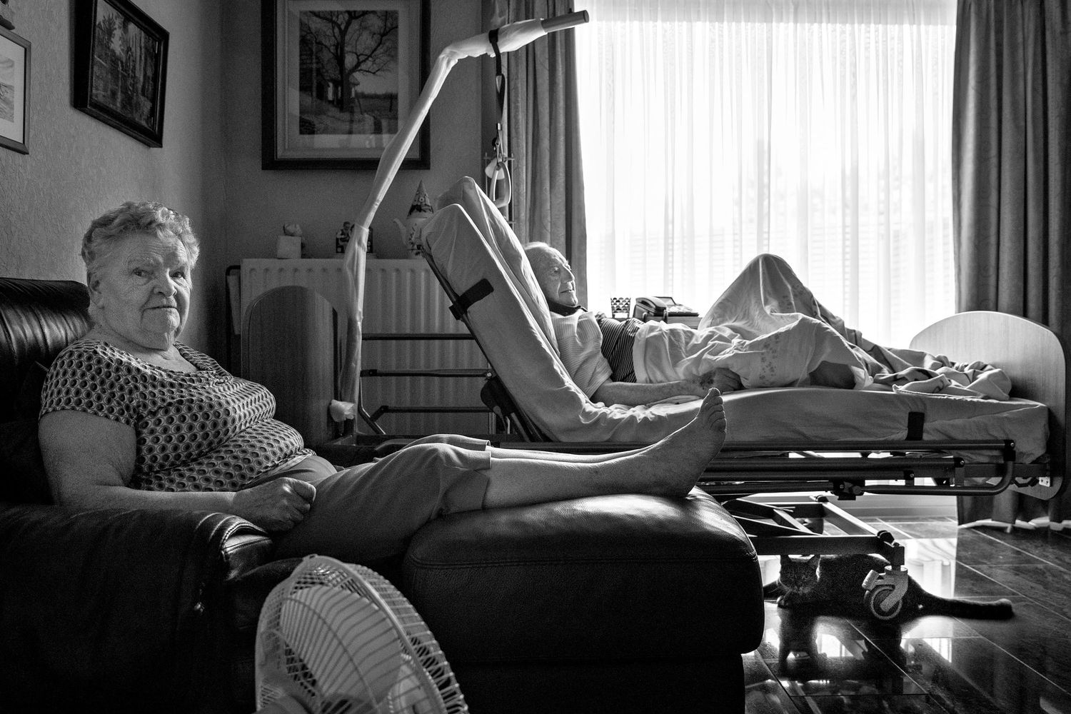 Black and white photo of elderly woman resting on sofa while another person exercises on a medical bed indoors.
