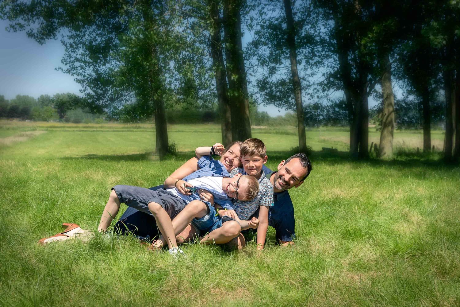 A group of people lying playfully in a grassy field with trees in the background on a sunny summer day.