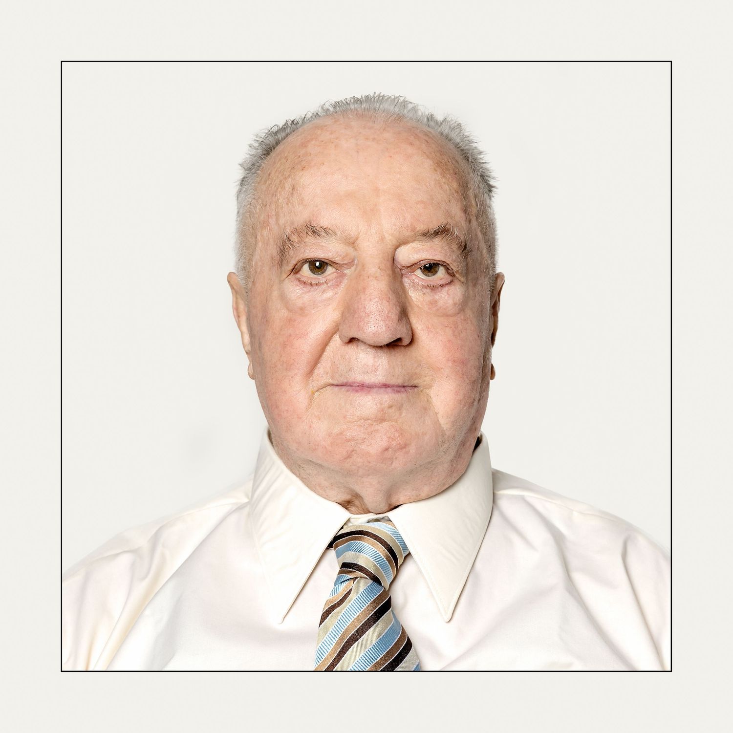 Elderly man in white dress shirt and striped tie poses for a formal portrait photo against a light background.