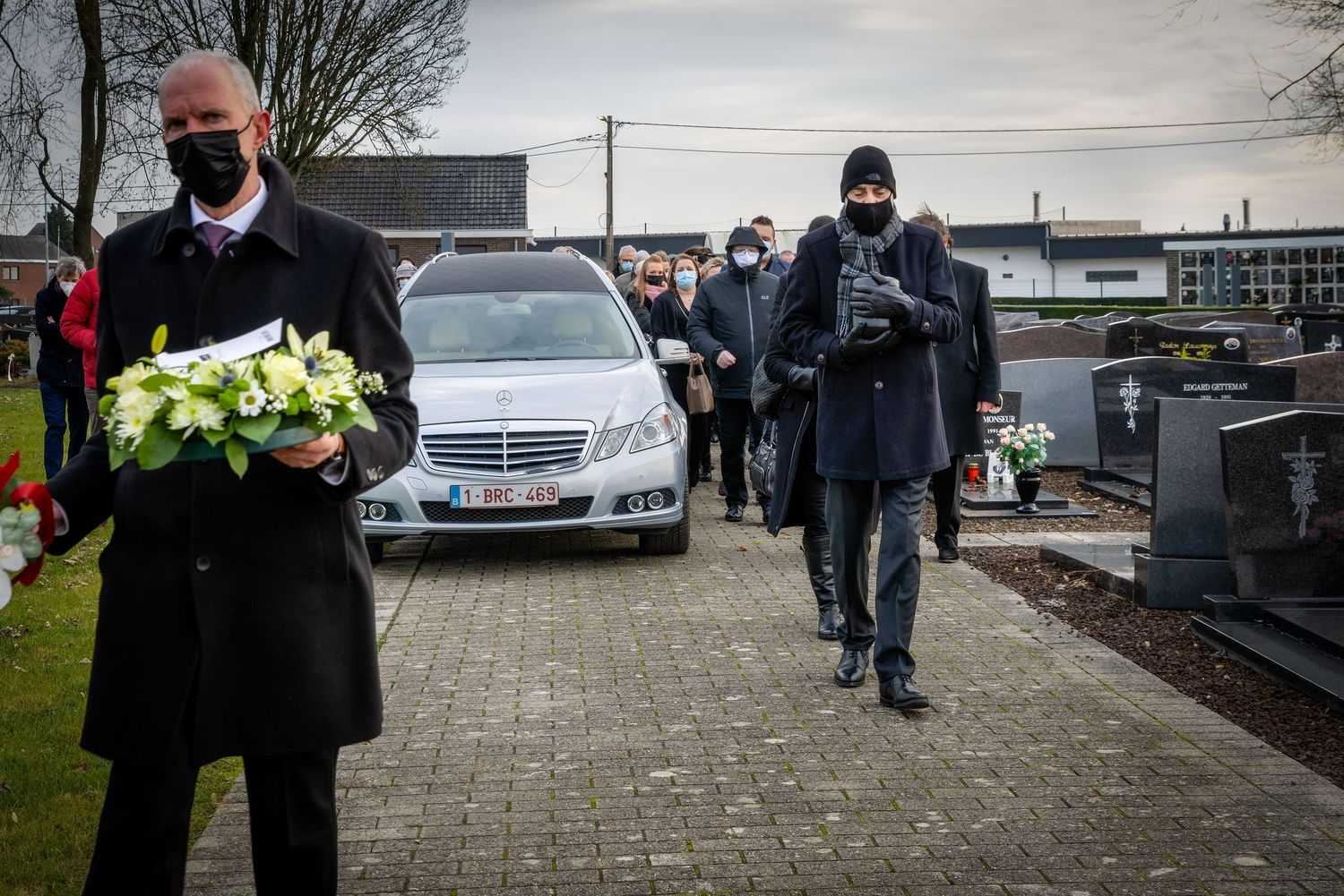 Mourners in black masks walk behind a hearse at a cemetery funeral service on a cloudy day.