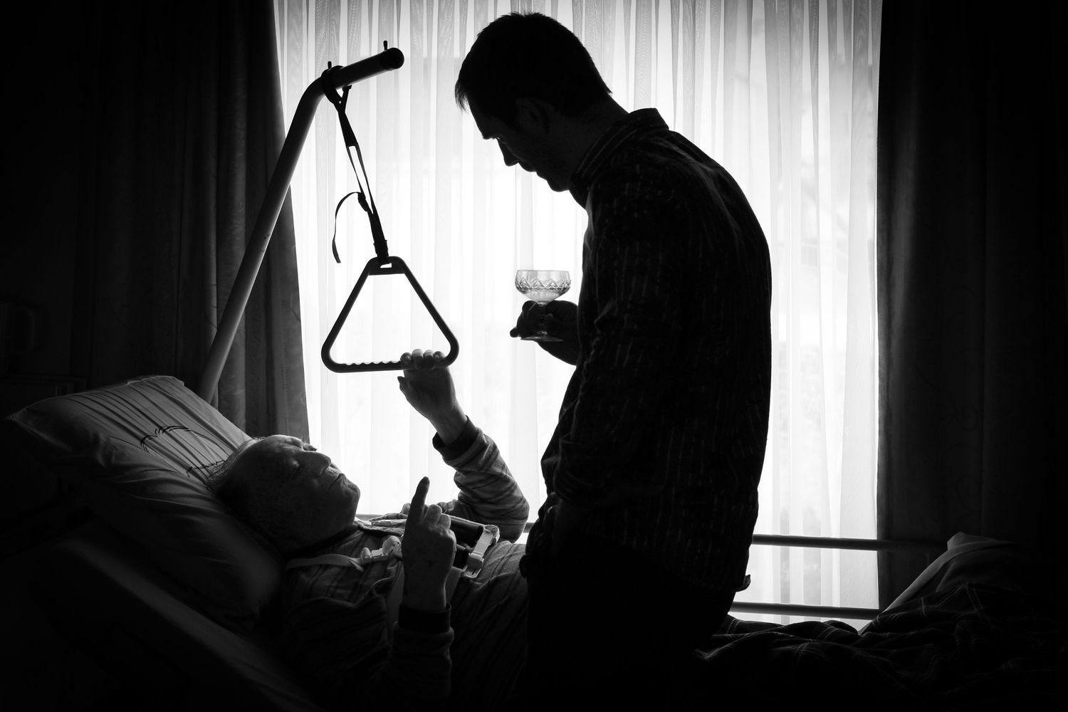 Black and white silhouette of caregiver tending to patient in hospital bed near window.