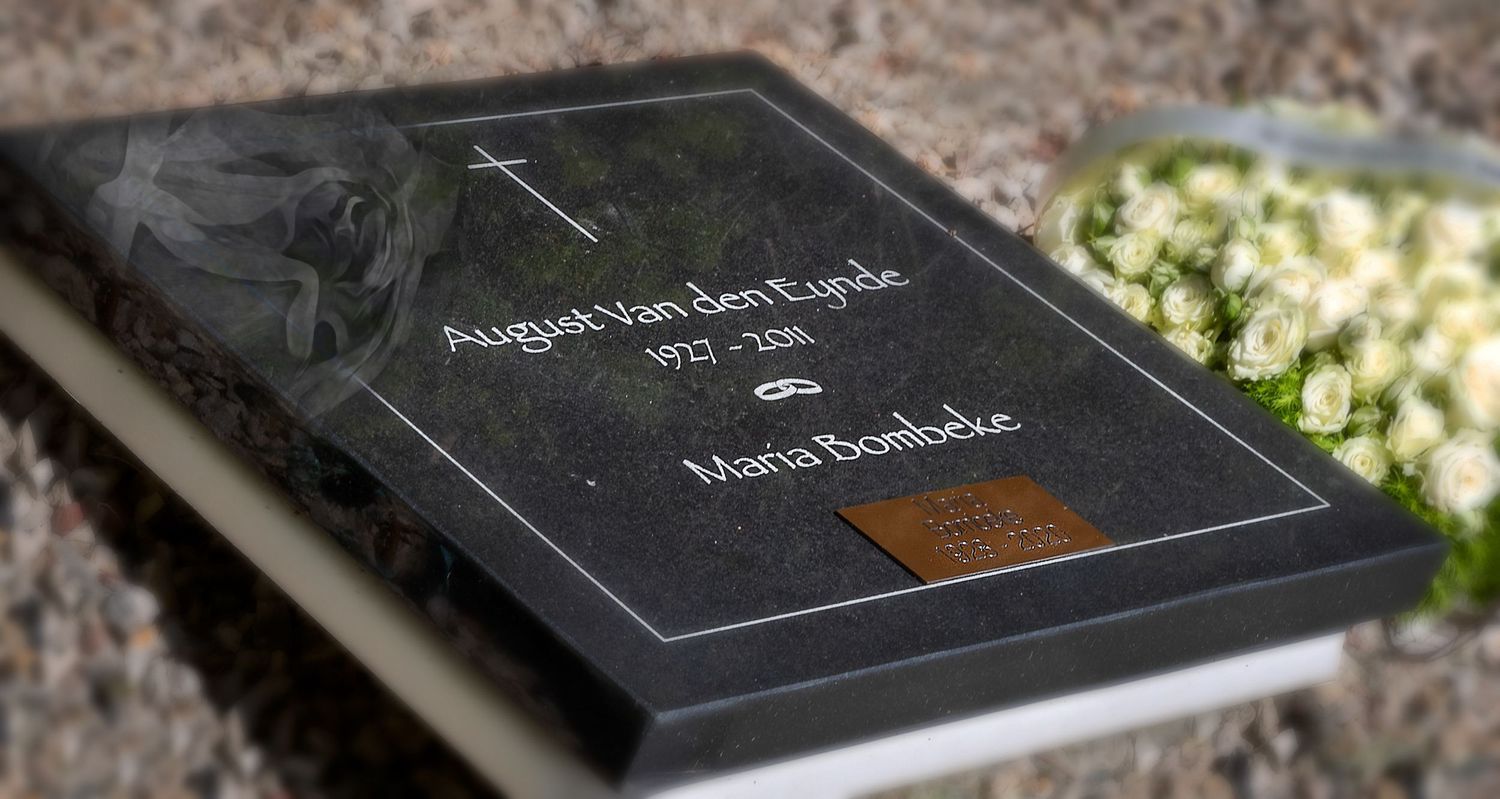 Dark granite memorial plaque for August Van den Eijnde and Maria Boelbeke with white flowers beside it.