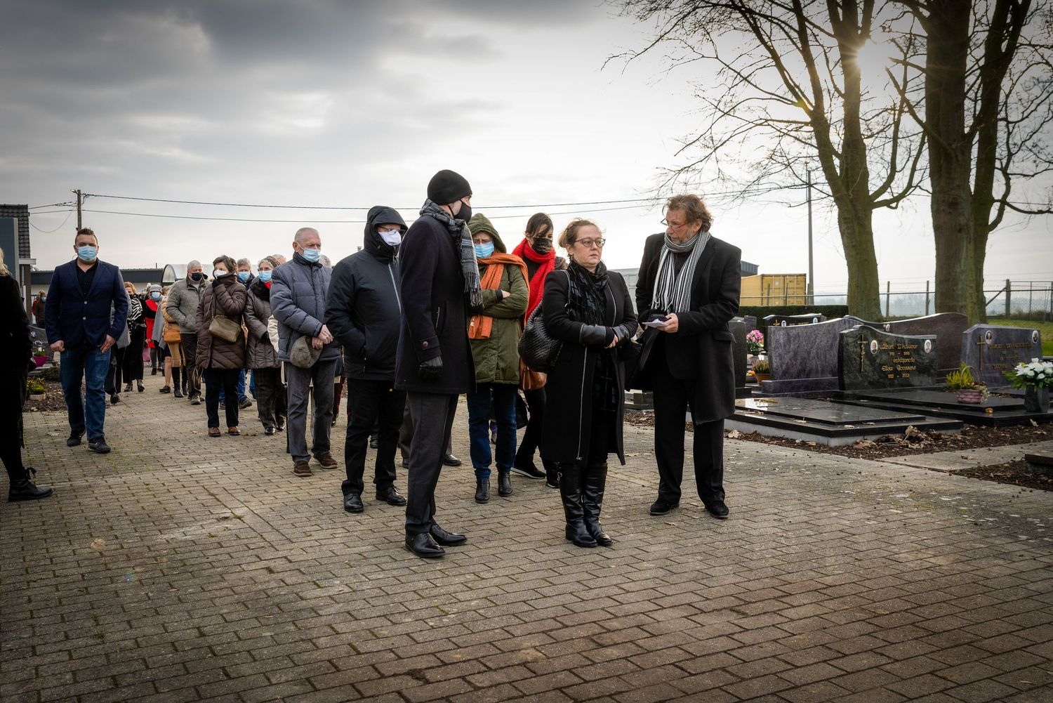 Group of people wearing masks walking in a line at an outdoor cemetery during a somber funeral gathering.