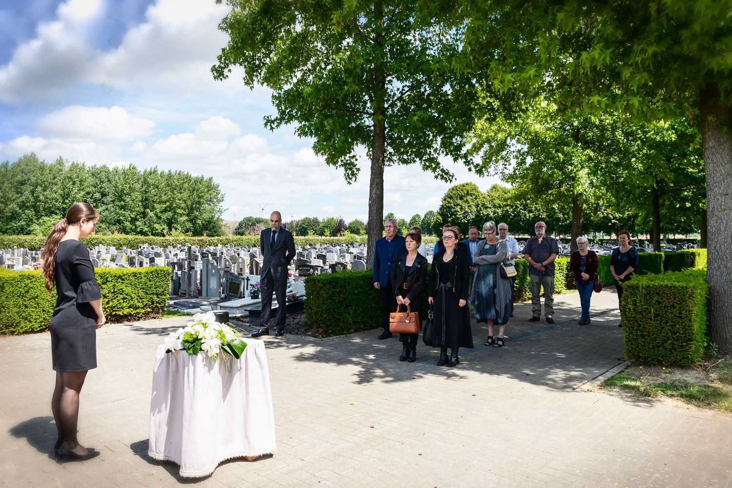 Outdoor funeral gathering at a cemetery with mourners dressed in black near a floral arrangement on a sunny day.