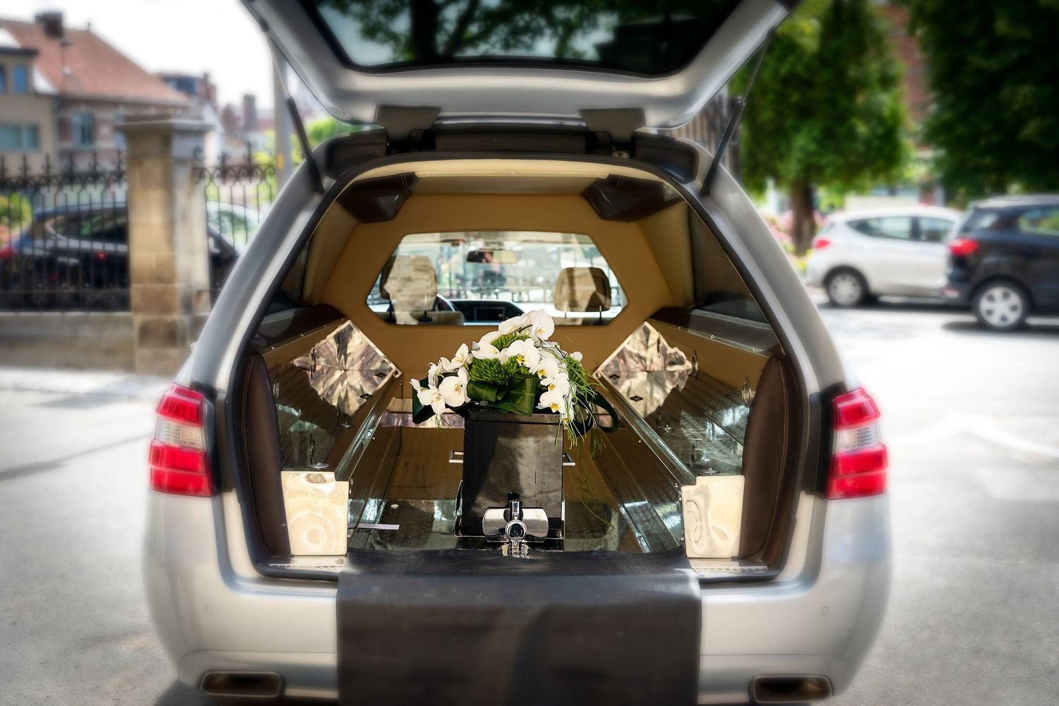 Hearse interior with golden casket handles and white floral arrangement visible through open rear door at funeral service.