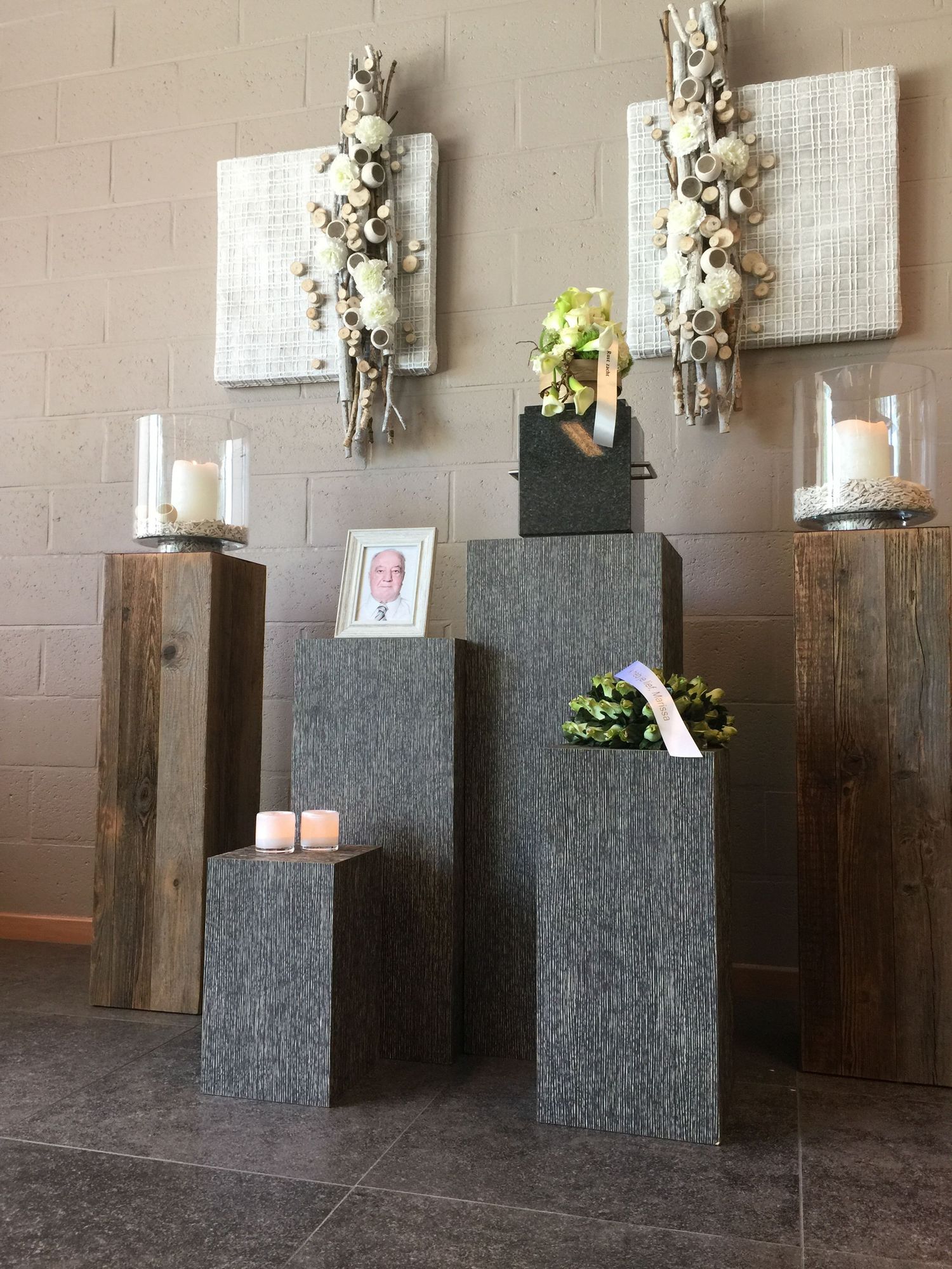 Funeral memorial display with gray pedestals, candles, framed photo, floral arrangements, and decorative wall art in a chapel.