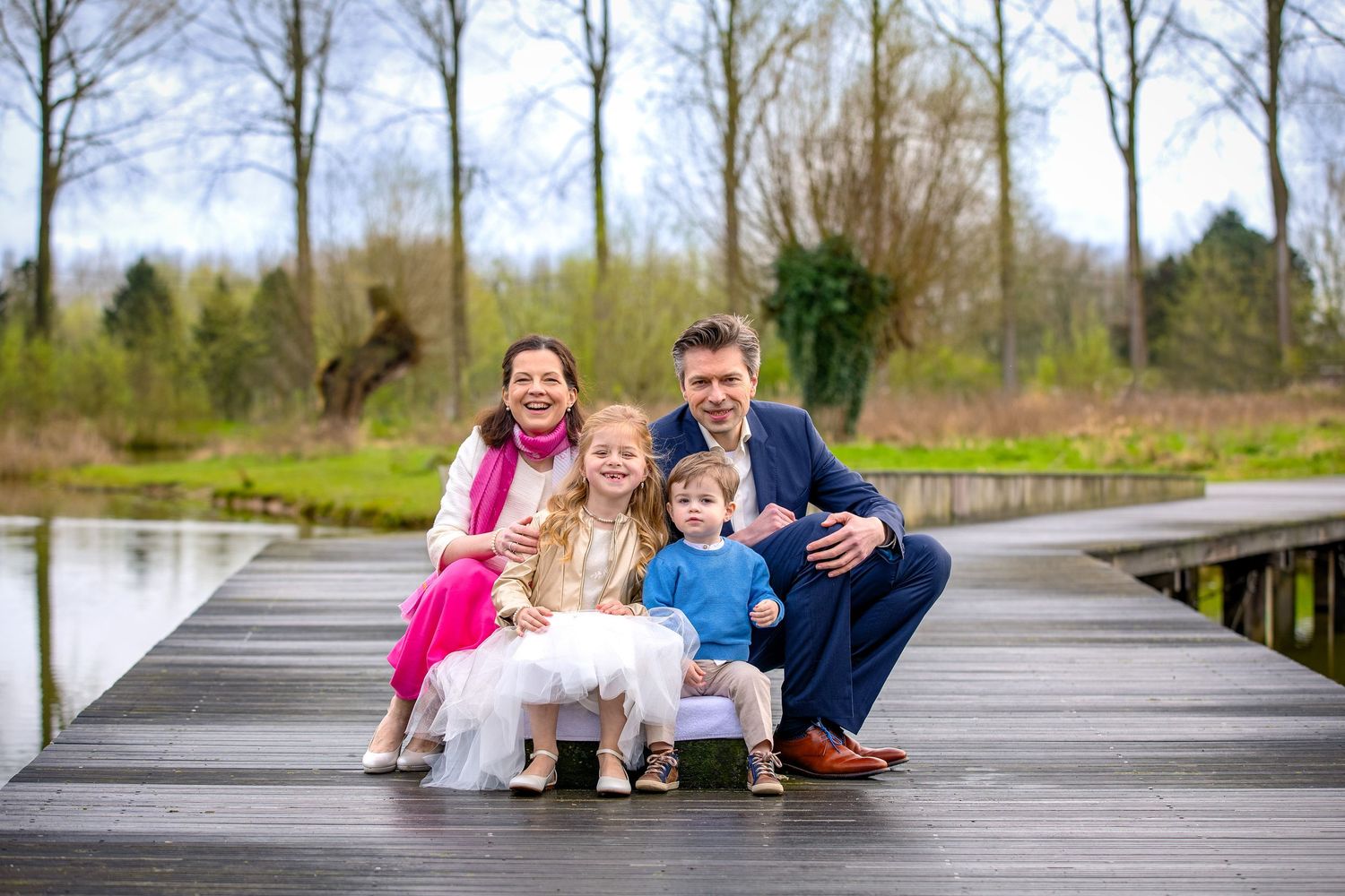 A family poses together on a wooden boardwalk over a pond surrounded by trees on a spring day.
