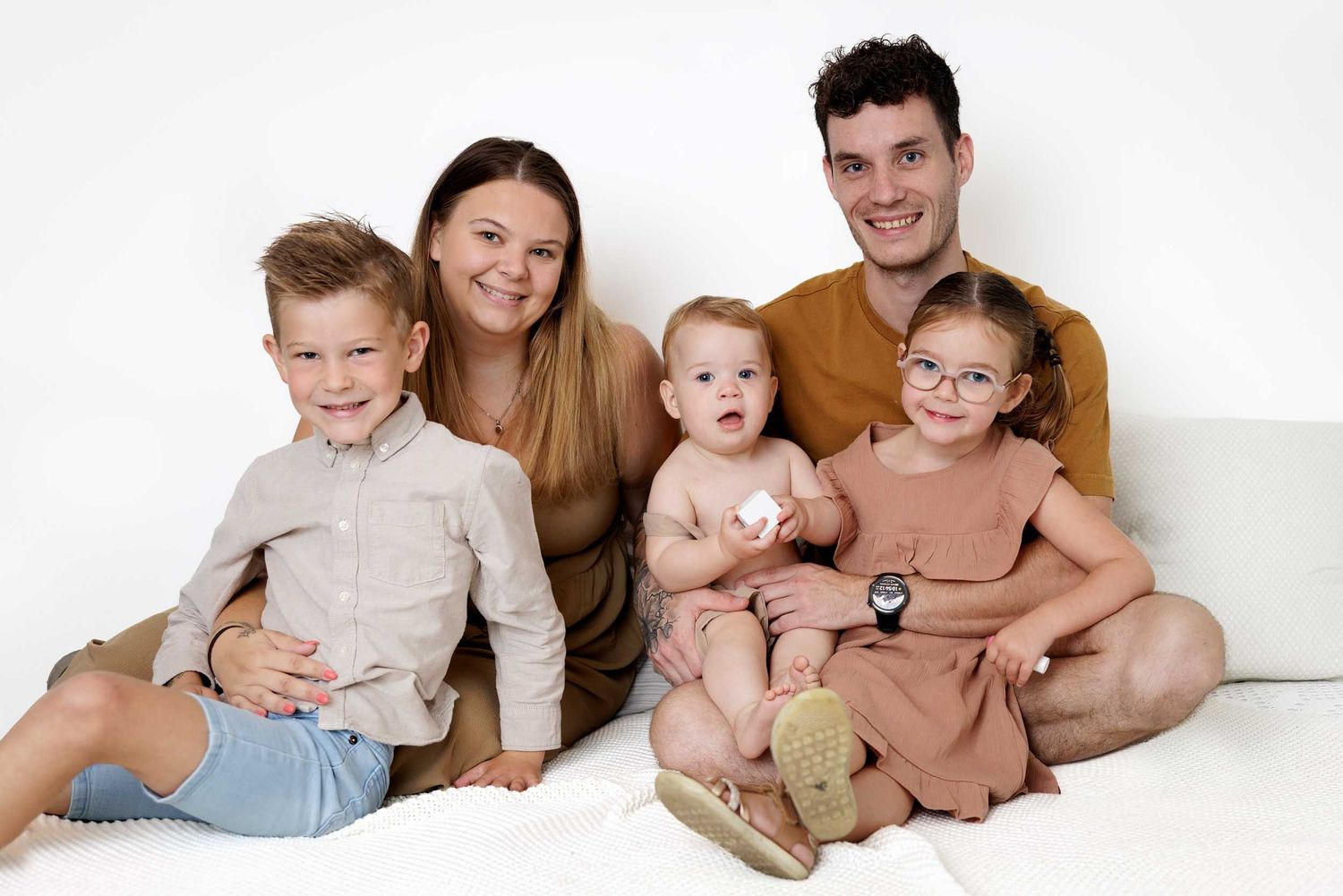 A happy family of five sits together on a white couch wearing neutral-colored casual clothing.