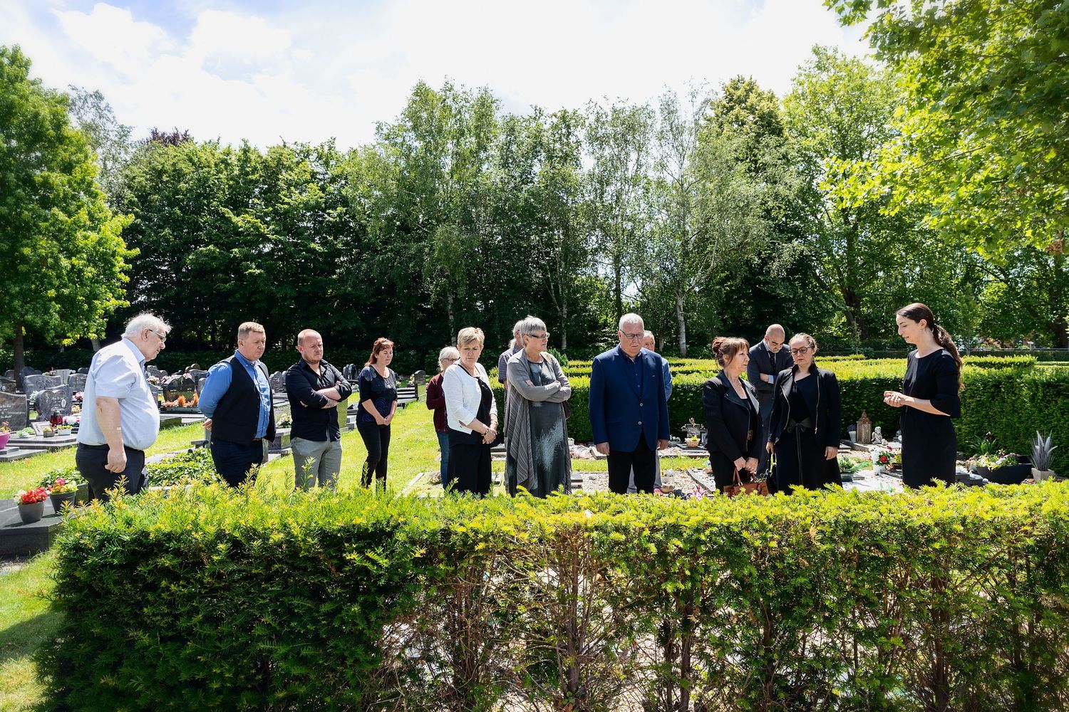 Group of people gathered at a cemetery memorial service surrounded by lush green trees and hedges on a sunny day.