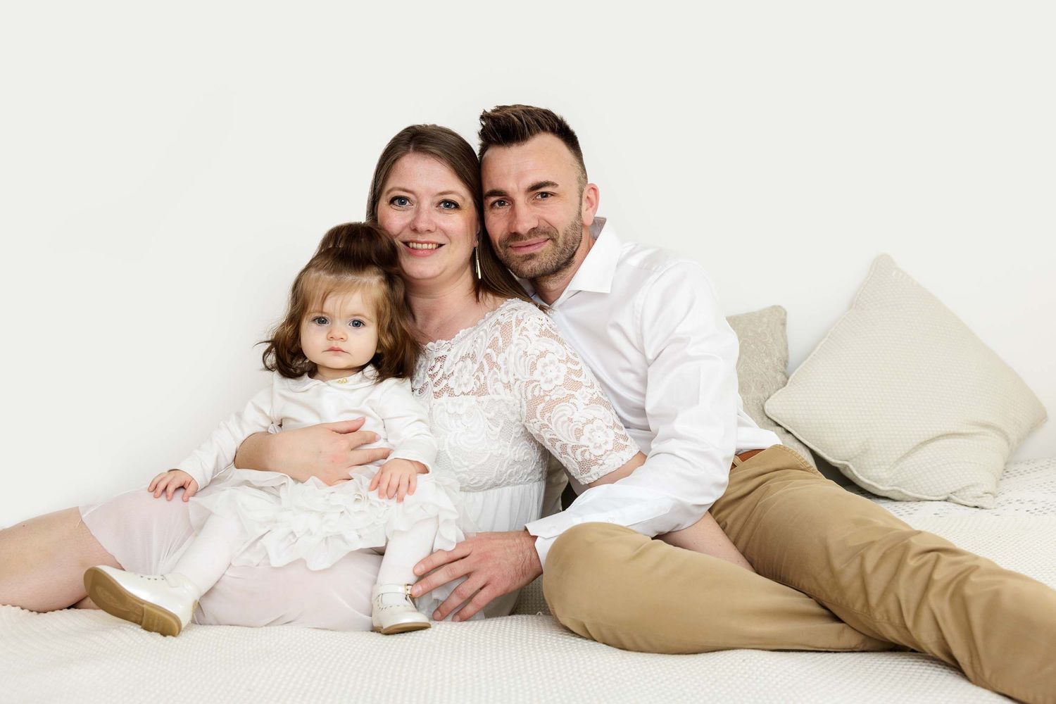 A family in coordinated white and beige outfits sits together on a light colored floor against a white background.