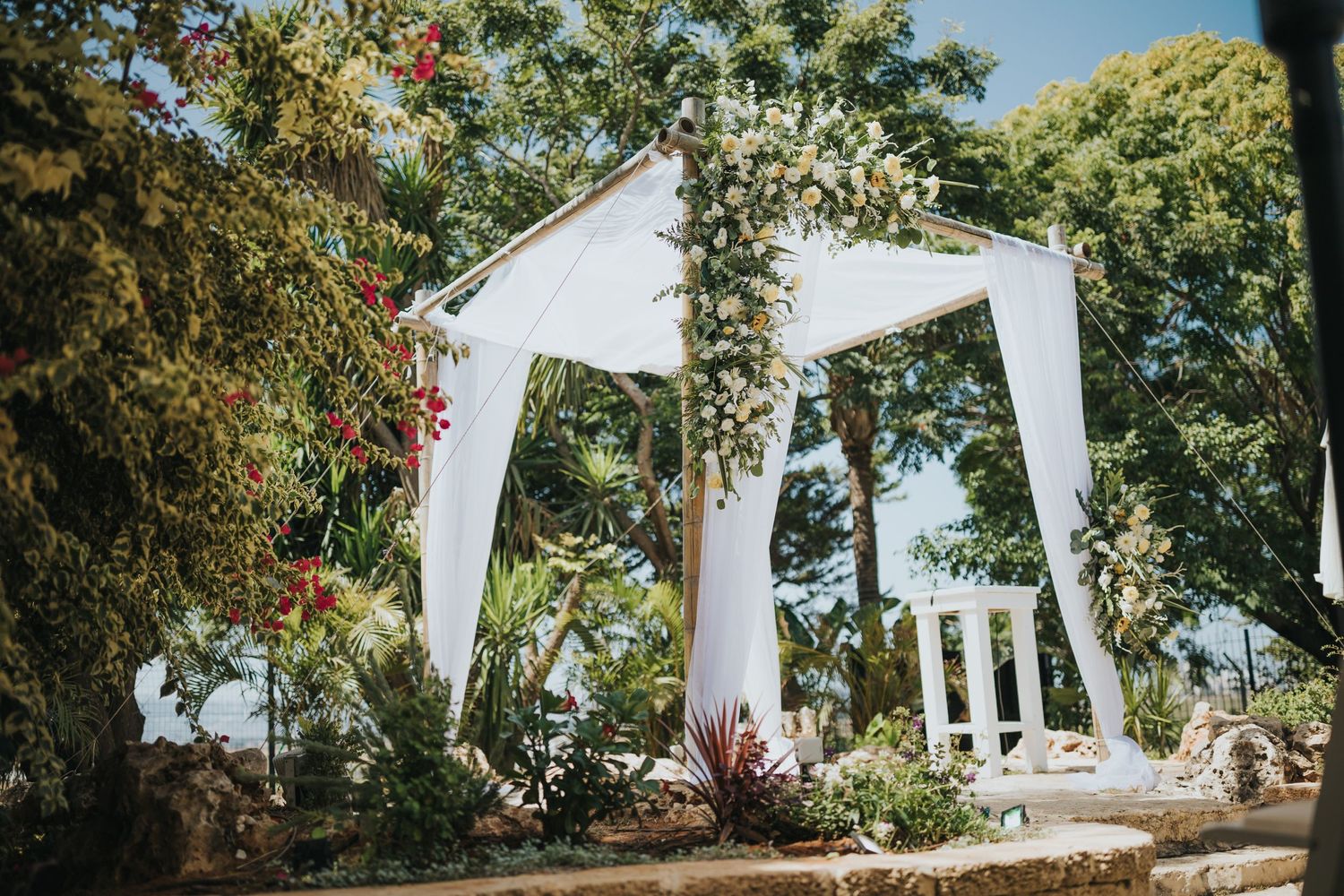 White wedding arch decorated with flowers and greenery against blue sky backdrop.