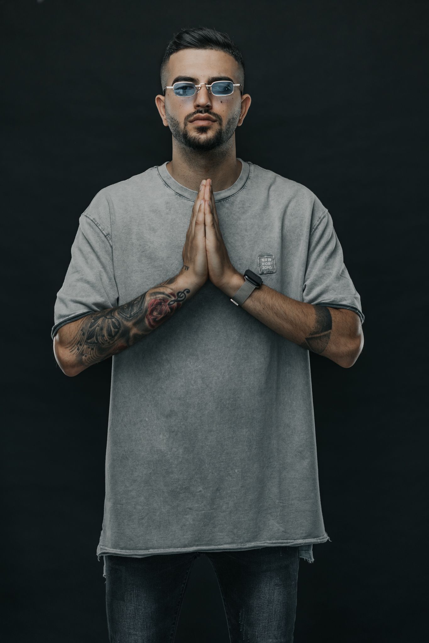 Person in grey t-shirt with tattoos and glasses holds hands in prayer position against dark background.