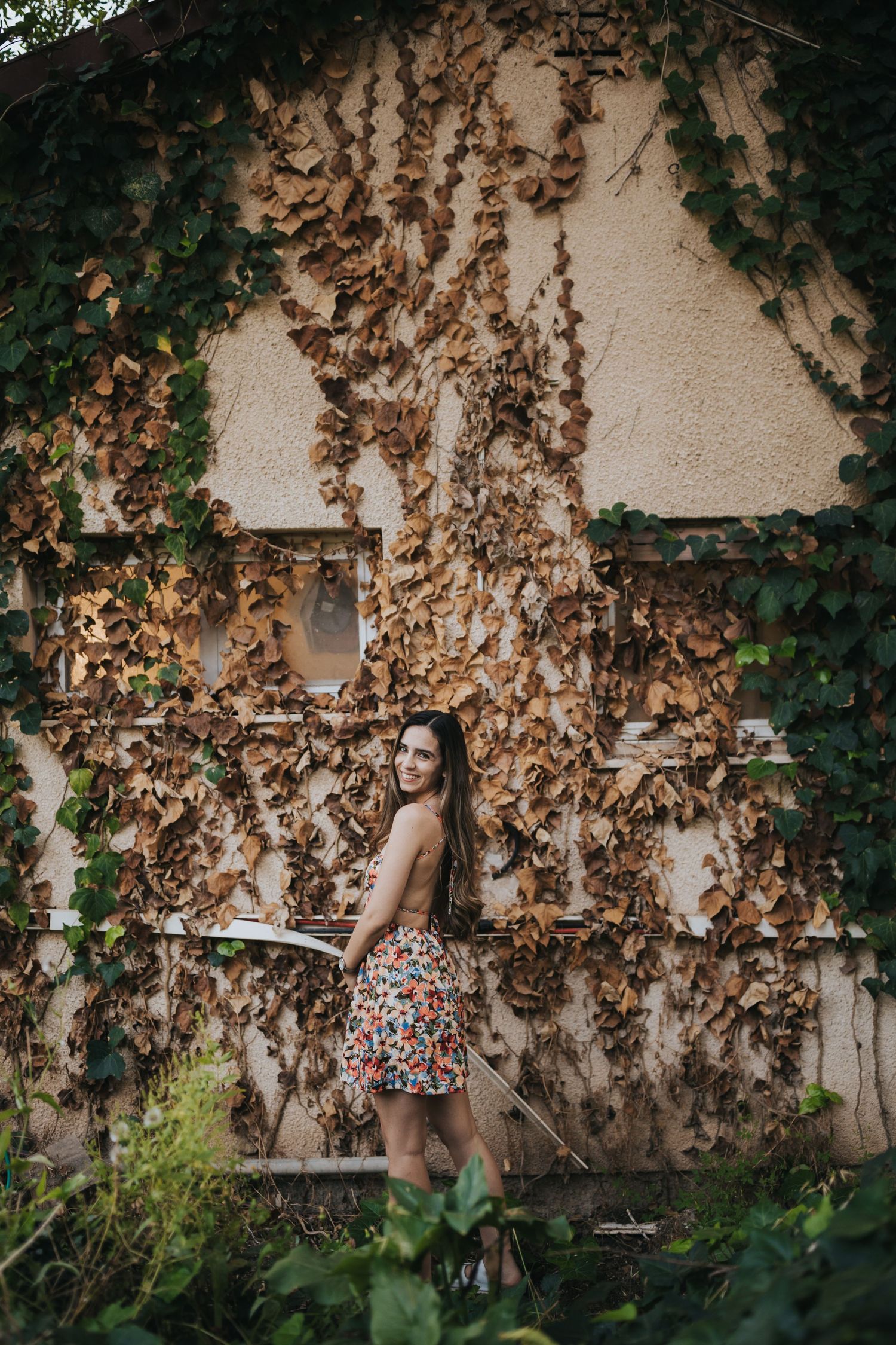 A vintage-inspired photo of someone posing against an ivy-covered wall in a floral dress.