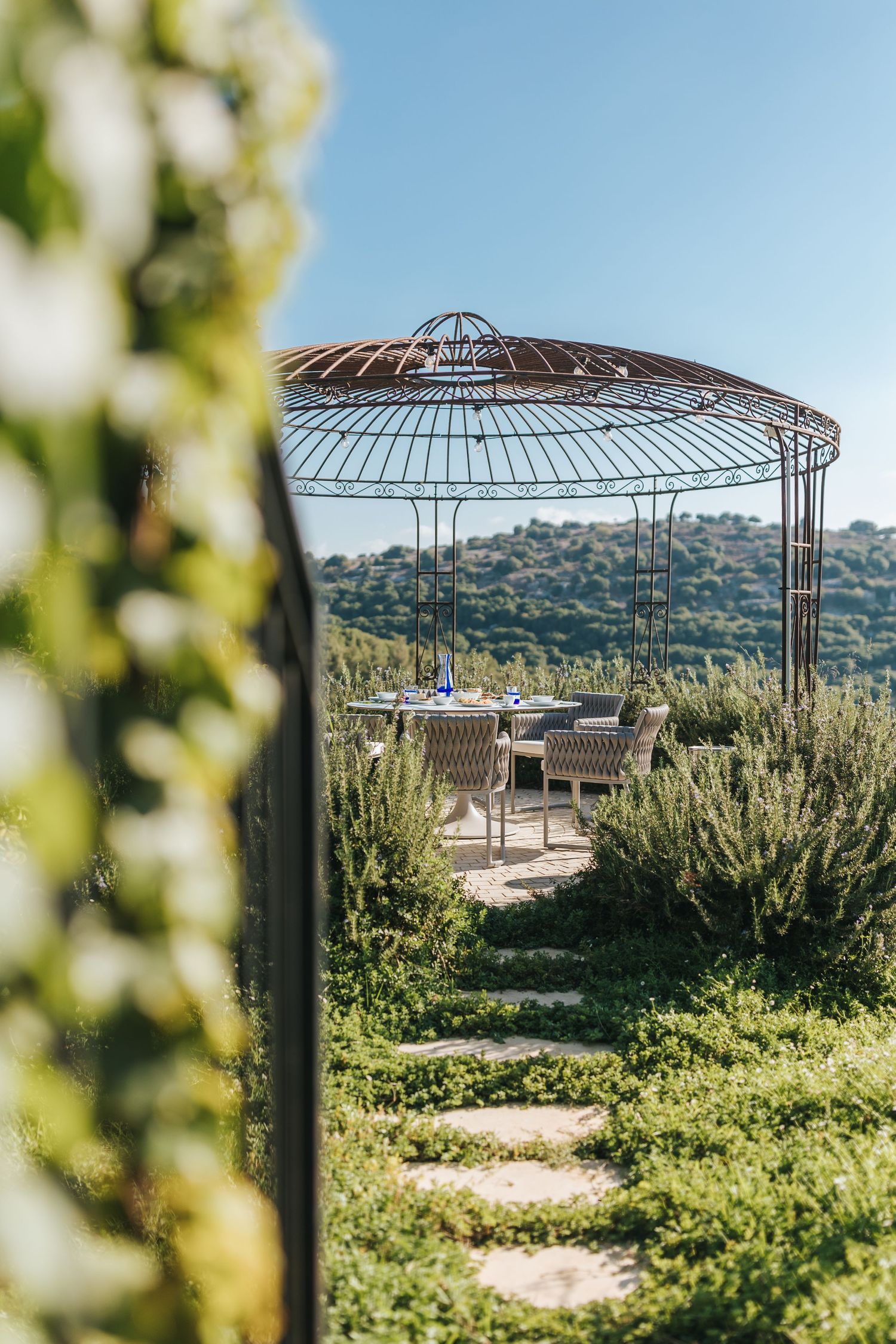Elegant metal gazebo with dining set overlooking scenic hillside landscape with lush greenery and blue sky.